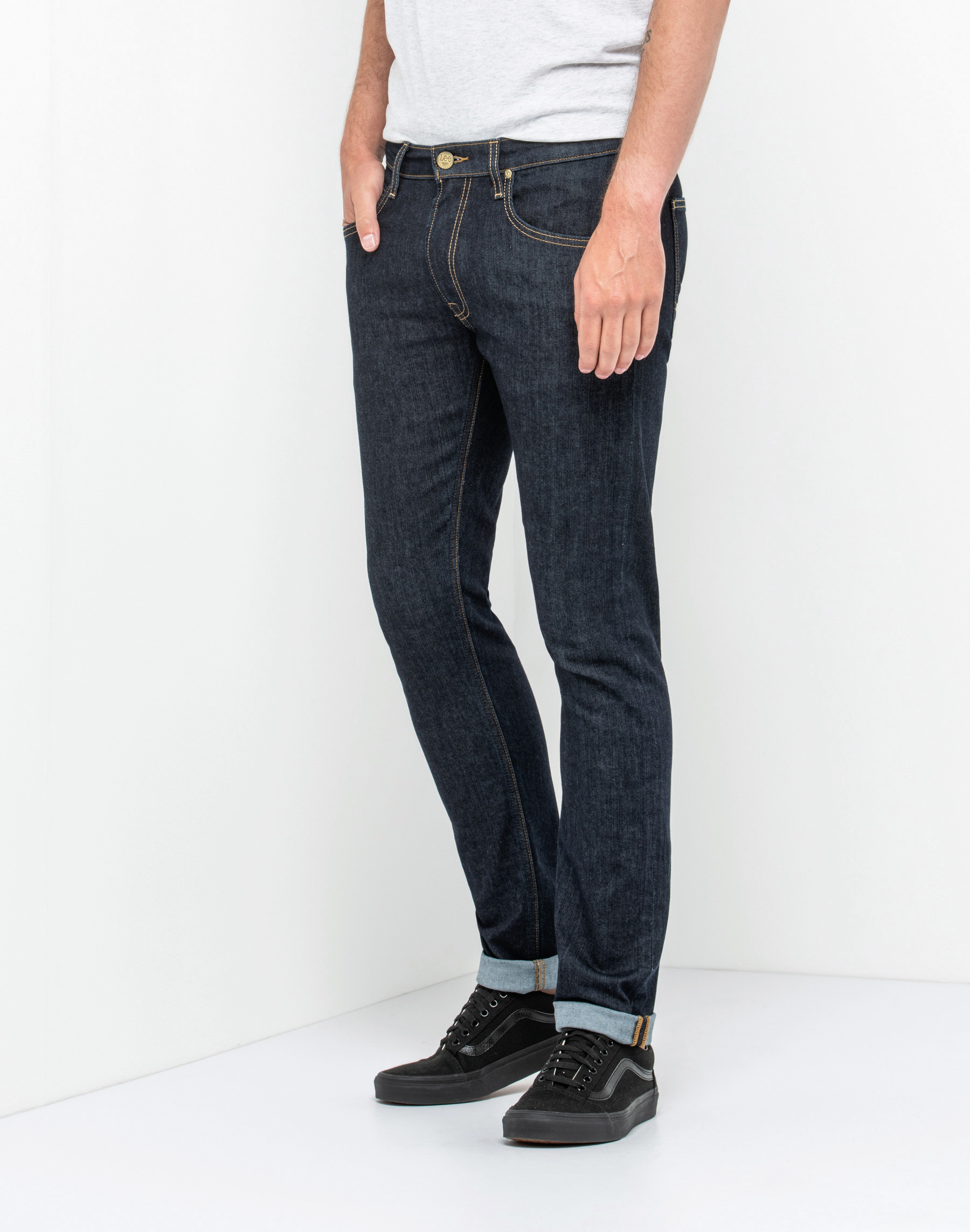 Pantalones vaqueros oscuros de corte recto con bajo vuelto y zapatillas negras sobre fondo blanco.
