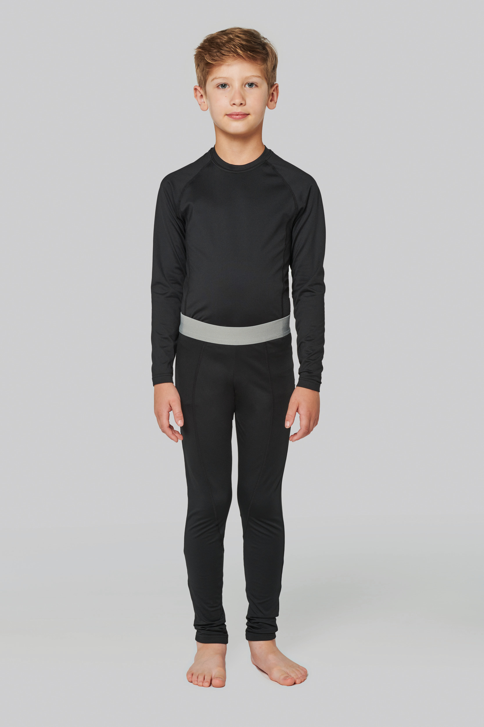 Niño de pie con camiseta y mallas térmicas negras de manga larga sobre fondo neutro.