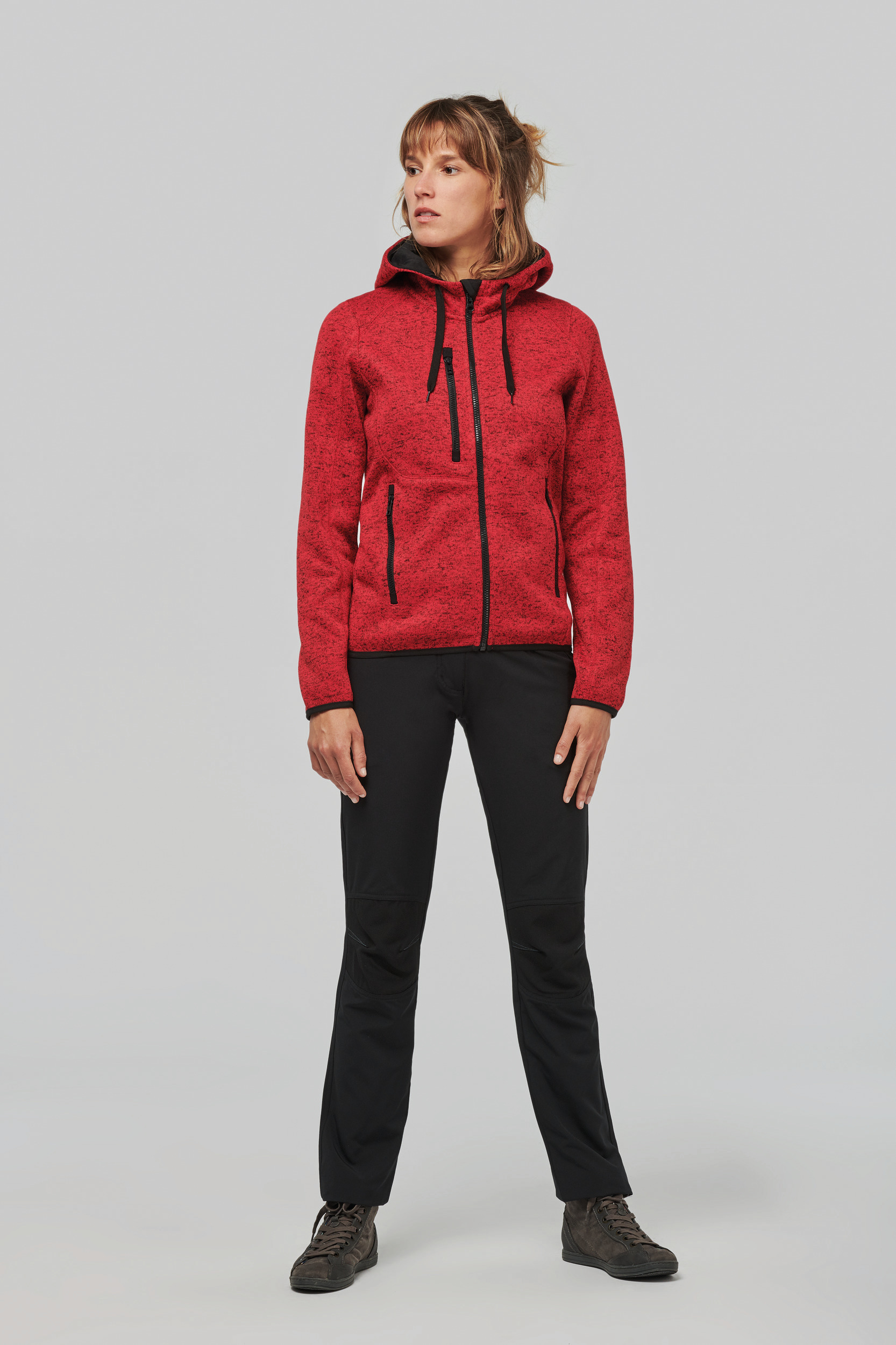 Mujer de pie con chaqueta roja de capucha y pantalones negros sobre fondo neutro