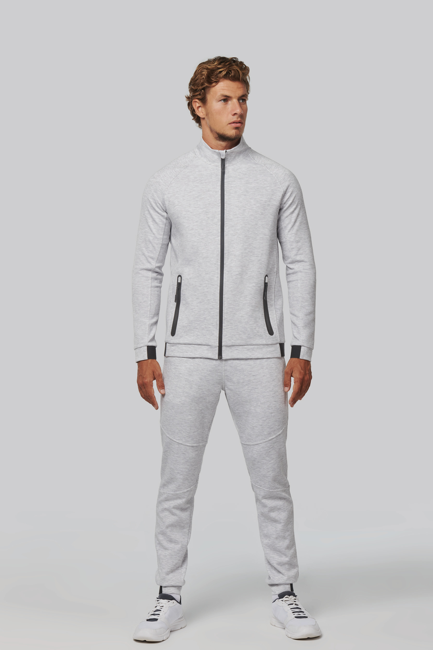 Hombre de pie con chándal gris claro con cremallera y bolsillos, luciendo zapatillas blancas sobre fondo neutro.