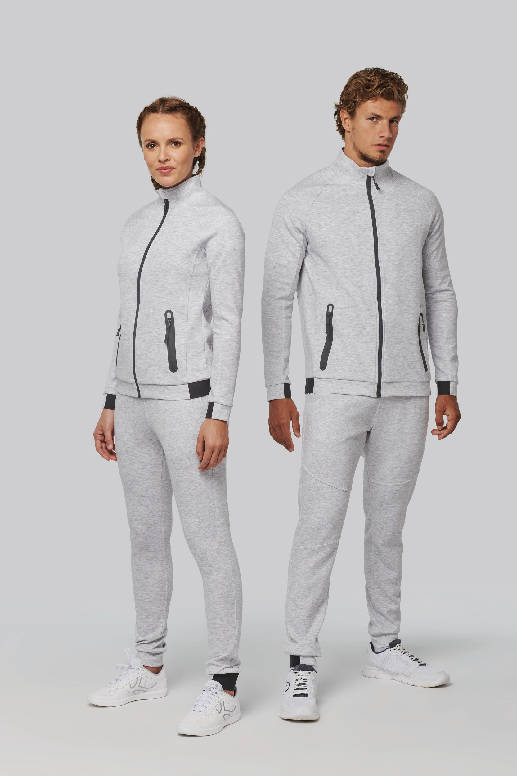 Hombre y mujer posando con chándal gris claro, chaqueta con cremallera y zapatillas blancas sobre fondo neutro