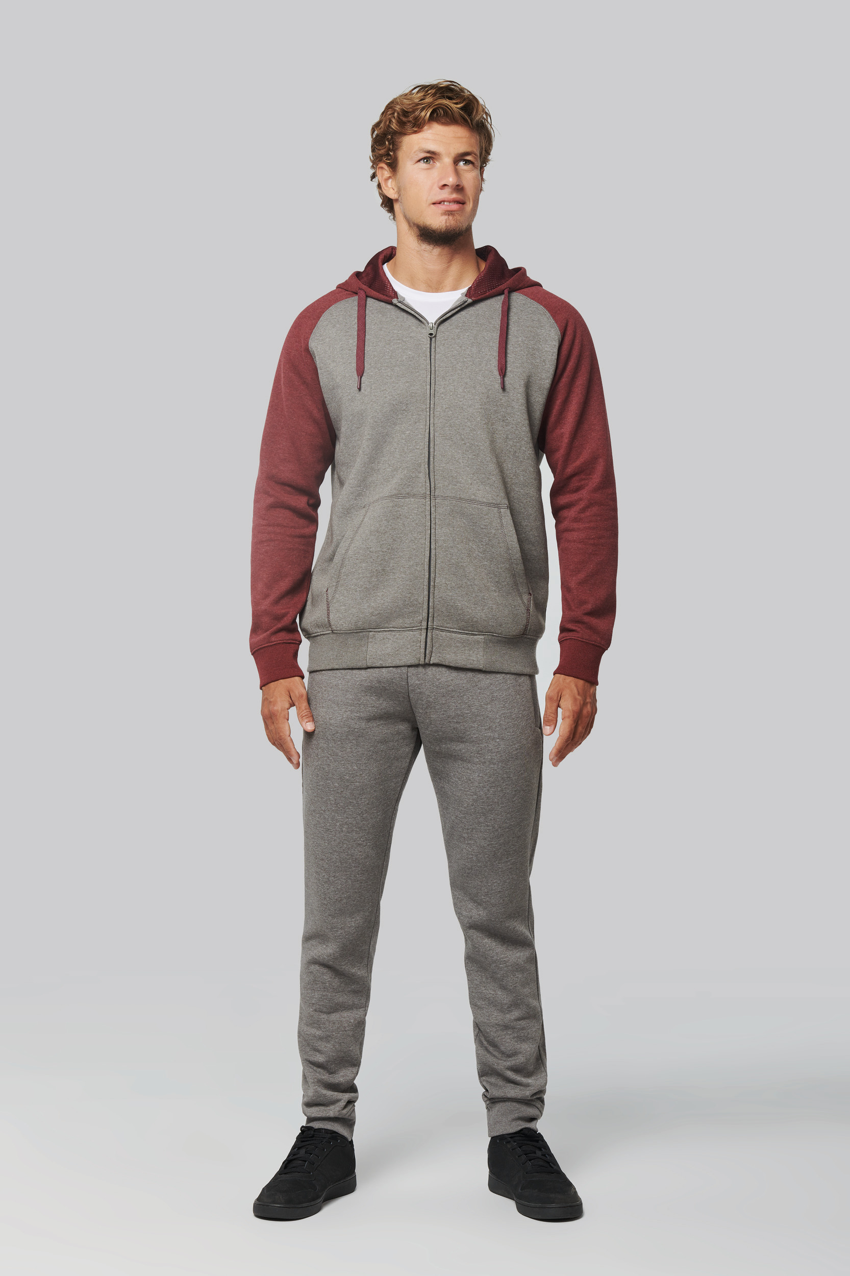 Hombre de pie con chándal gris y capucha granate sobre fondo neutro