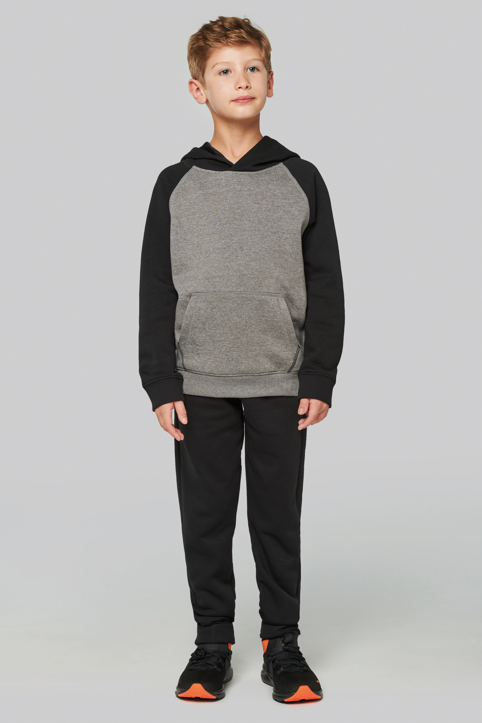 Niño de pie con sudadera gris y negra y pantalón deportivo negro en posado sobre fondo claro