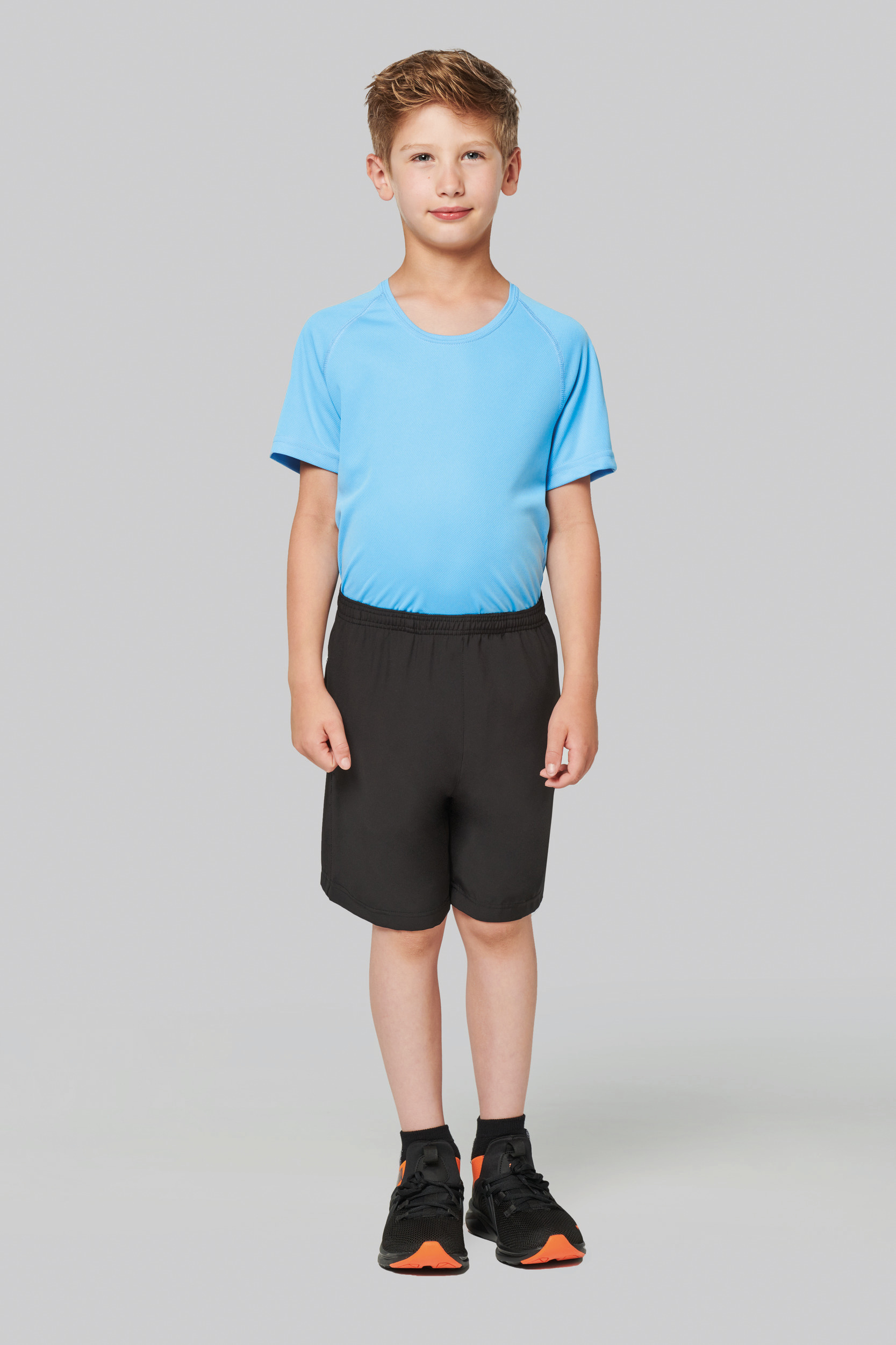 Niño de pie con camiseta deportiva azul y pantalones cortos negros sobre fondo gris