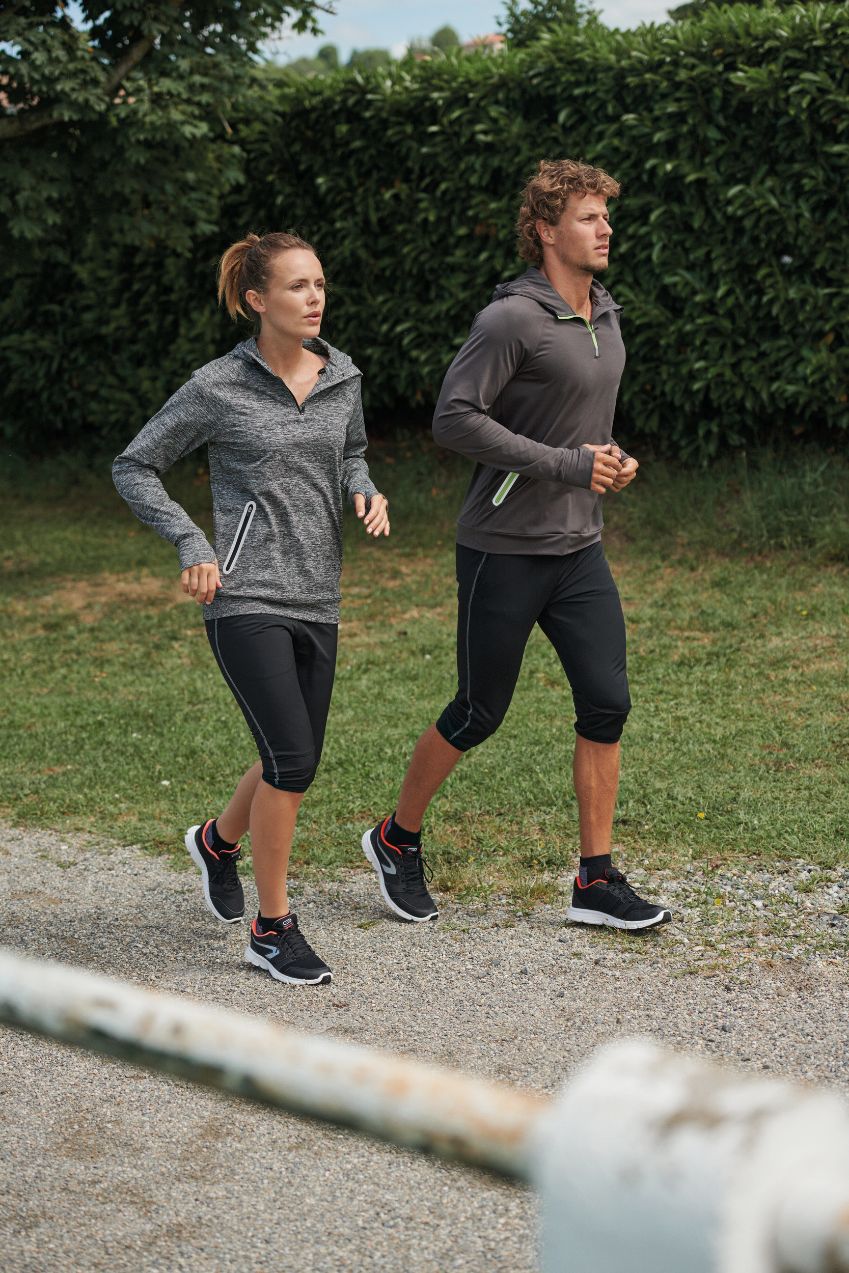 Hombre y mujer corriendo al aire libre por un camino de grava junto a setos verde.