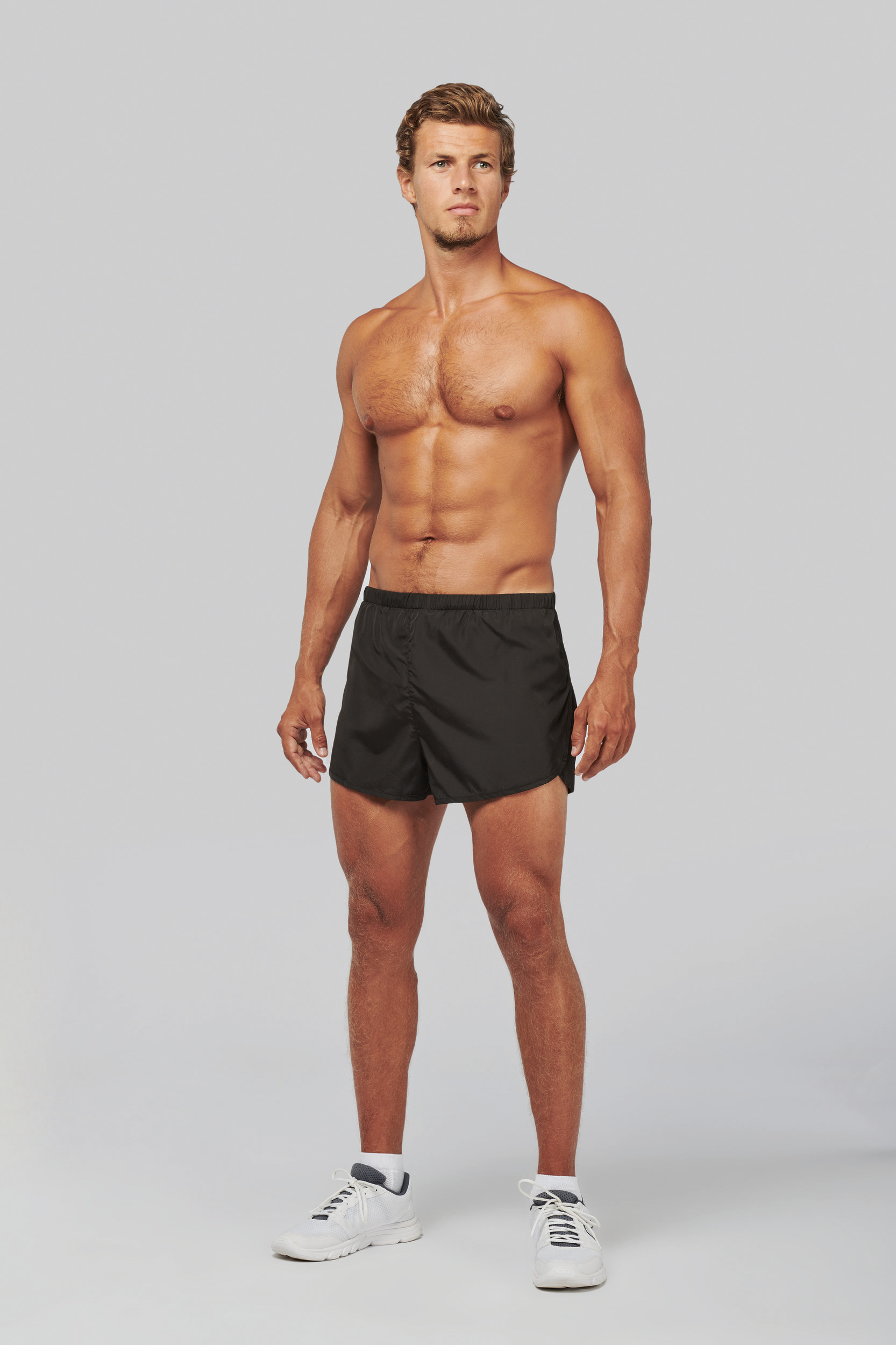 Hombre atlético sin camiseta con pantalones cortos negros de deporte y zapatillas blancas sobre fondo gris