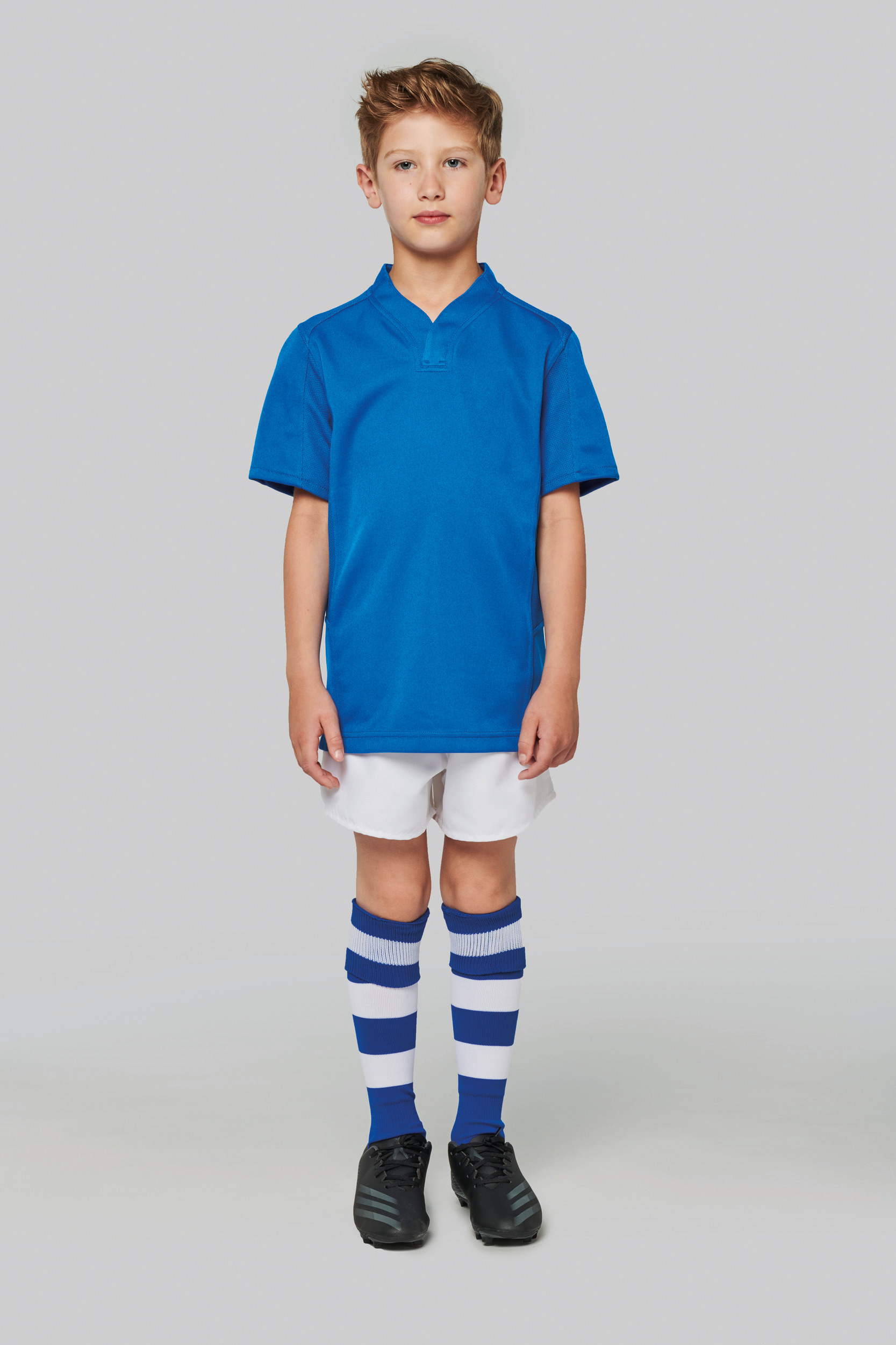 Niño con camiseta azul de fútbol, pantalón blanco, calcetas a rayas azules y blancas y botas negras sobre fondo neutro