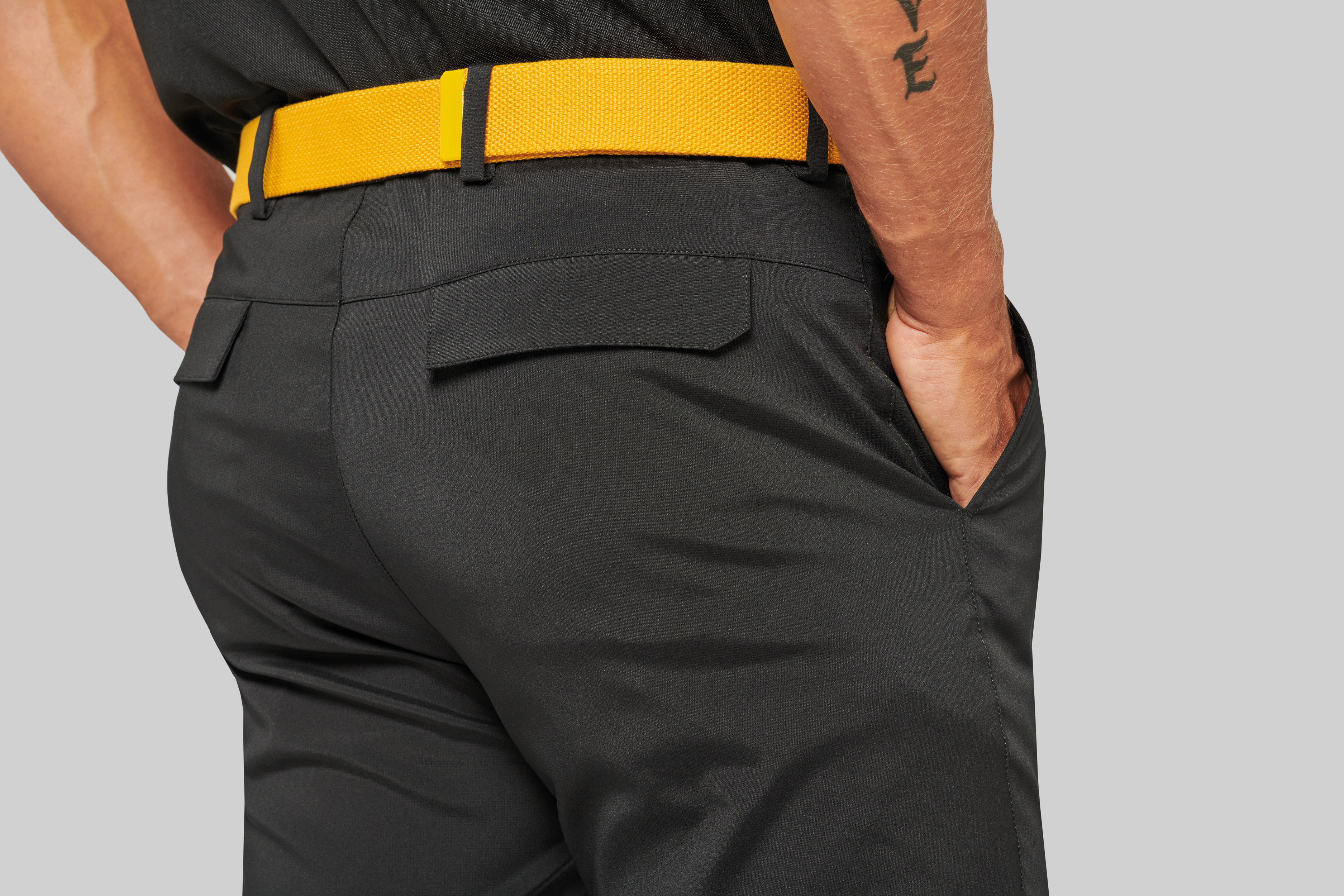 Hombre de espaldas con pantalones negros, cinturón amarillo y mano en el bolsillo delantero.