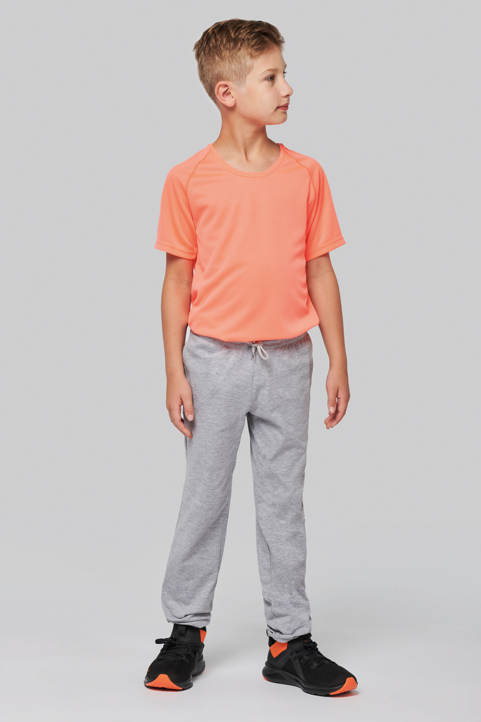 niño con camiseta deportiva naranja y pantalones grises de chándal de perfil sobre fondo neutro