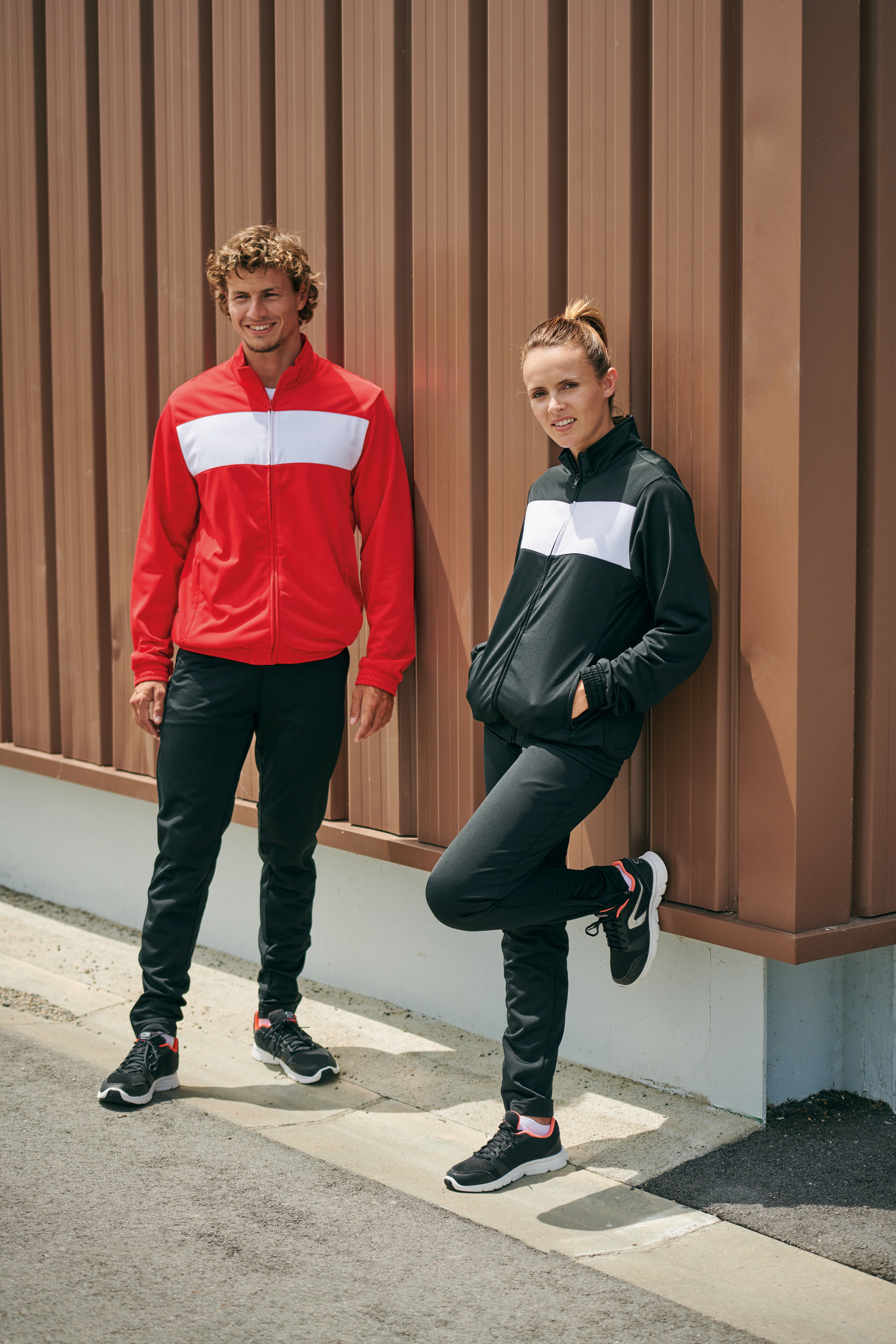 Pareja apoyada en una pared de metal vistiendo chándal deportivo con franjas blancas