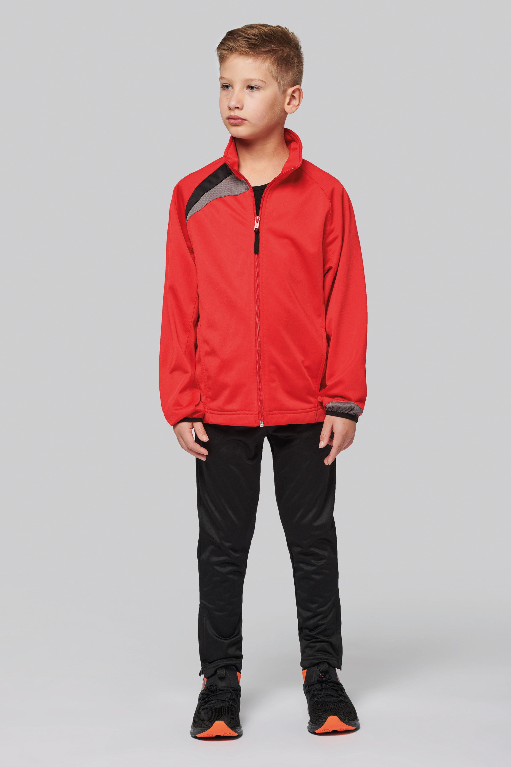 Niño de pie con chándal rojo y pantalones negros sobre fondo neutro.