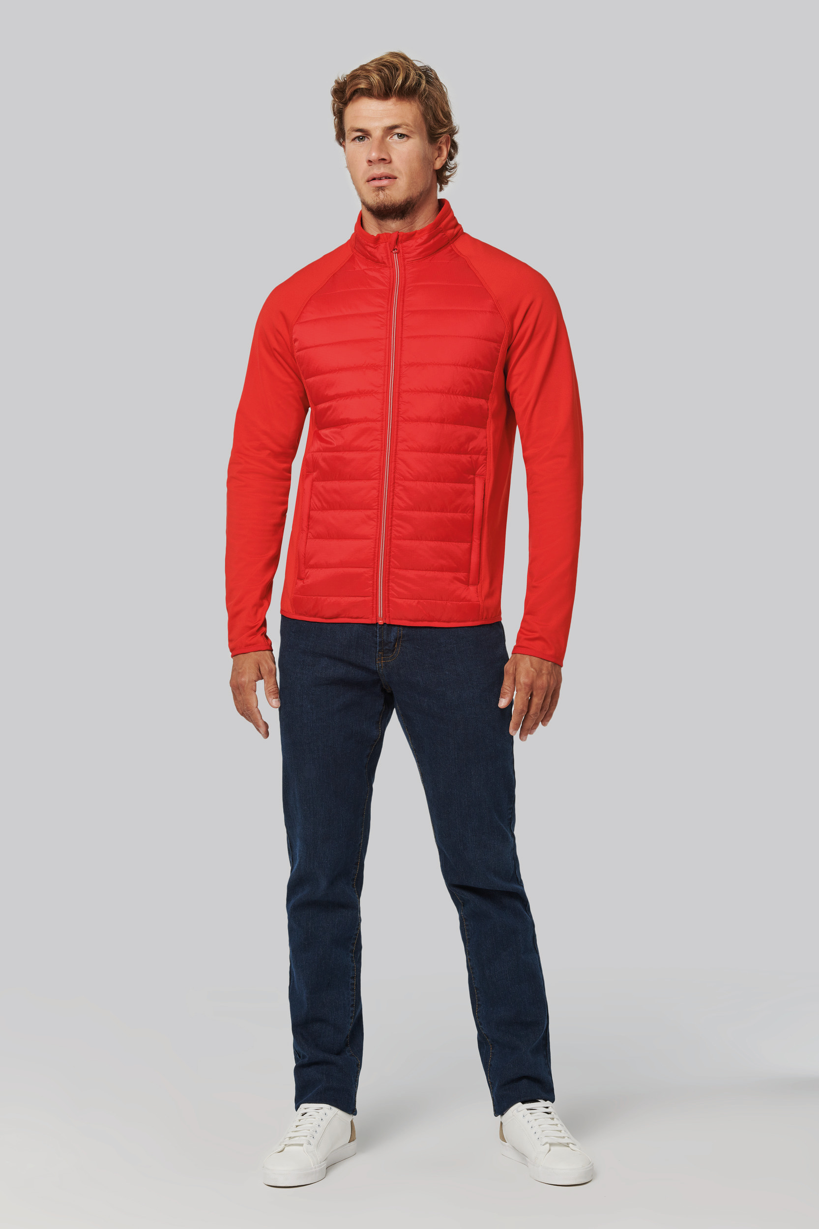 Hombre de pie con chaqueta roja acolchada, vaqueros y zapatillas blancas sobre fondo gris.