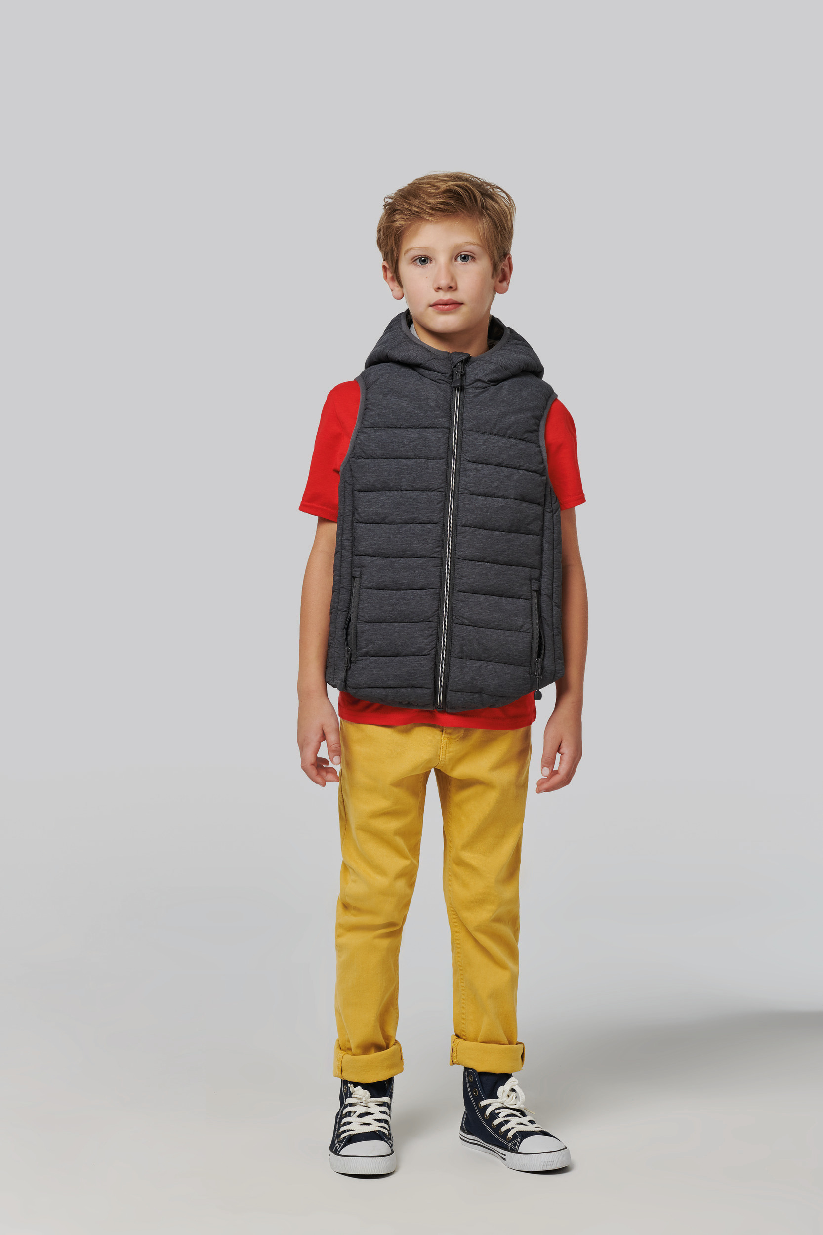 Niño de pie con chaleco acolchado gris y capucha, camiseta roja, pantalones amarillos y zapatillas de lona azul frente a fondo neutro.