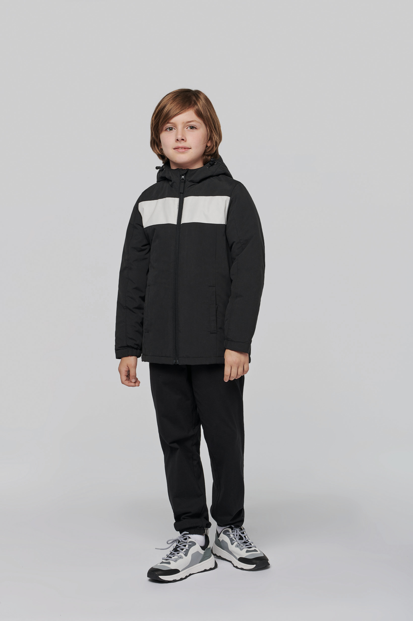 Niño de pie con cazadora acolchada negra con franja blanca, pantalones oscuros y zapatillas deportivas grises sobre fondo gris.