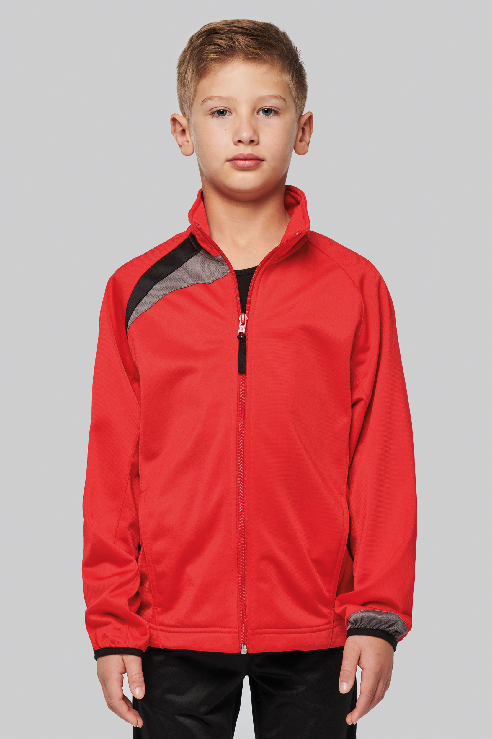 Niño de pie con chaqueta deportiva roja de cremallera y detalle negro sobre fondo neutro.