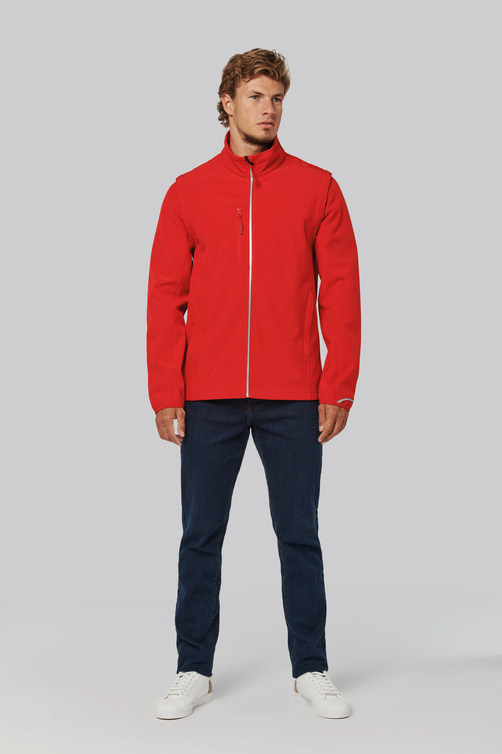 Hombre con chaqueta softshell roja de cremallera y bolsillo en el pecho, vaqueros oscuros y zapatillas blancas sobre fondo gris.
