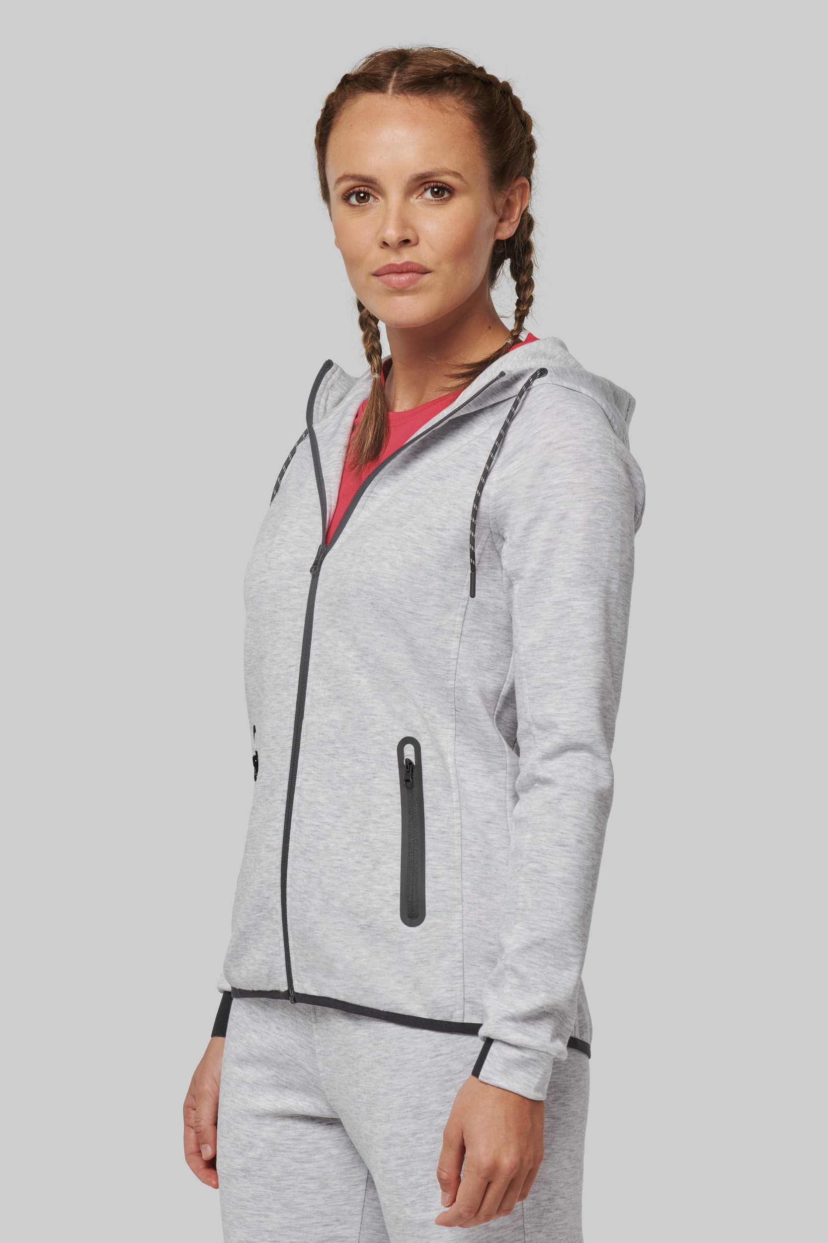 Mujer joven con sudadera gris con capucha y pantalón deportivo posando de pie sobre fondo neutro