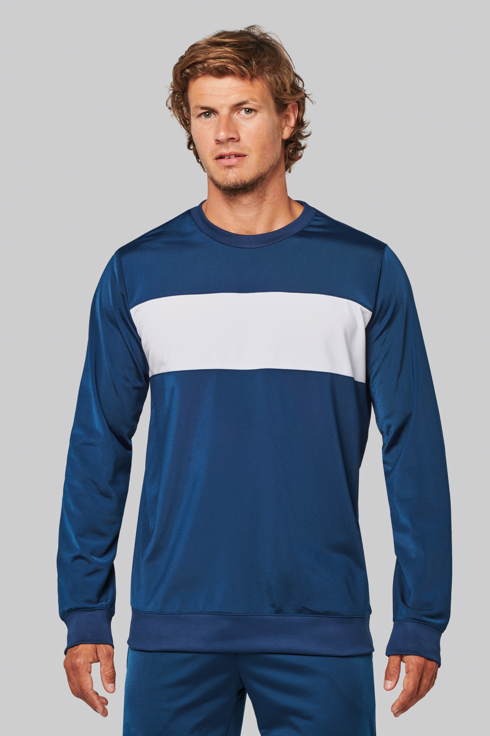 hombre de pie con sudadera azul de manga larga y franja horizontal blanca sobre fondo neutro