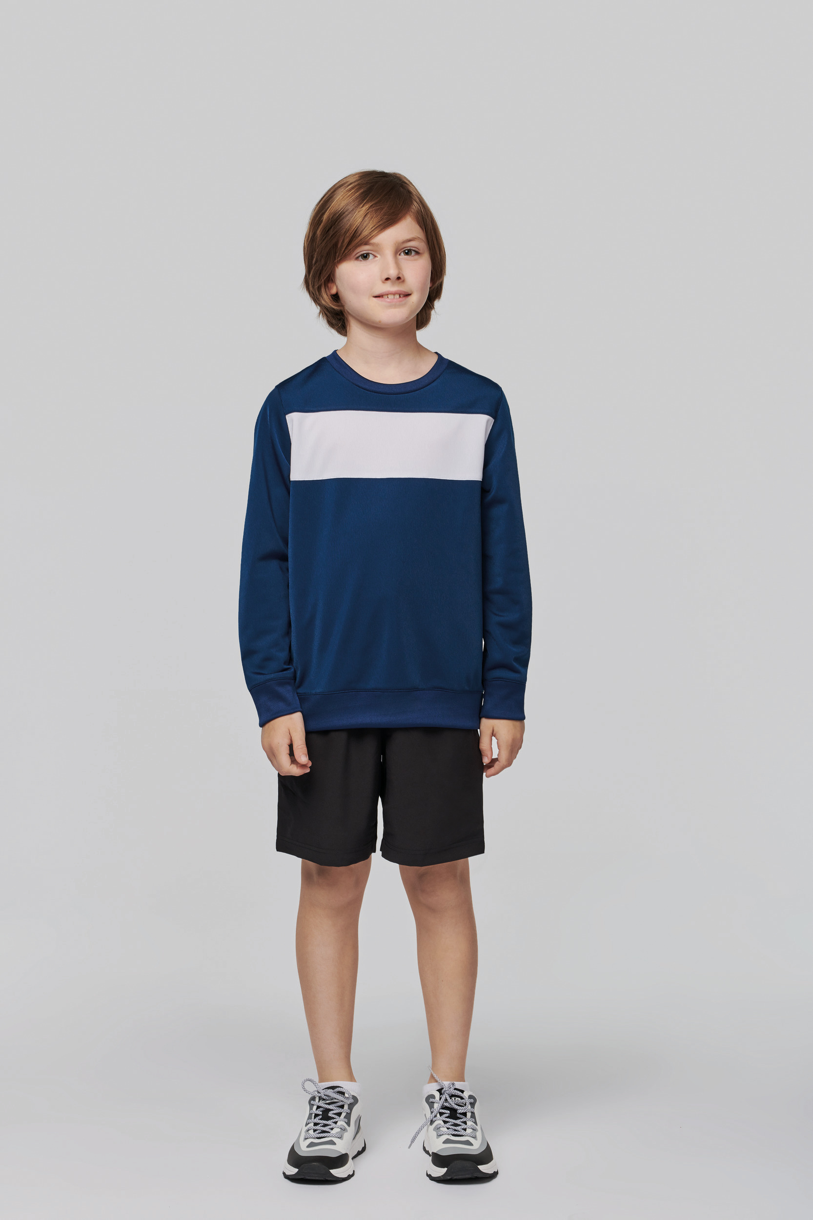 Niño de pie con sudadera azul con franja blanca, pantalones cortos negros y zapatillas deportivas sobre fondo gris claro.