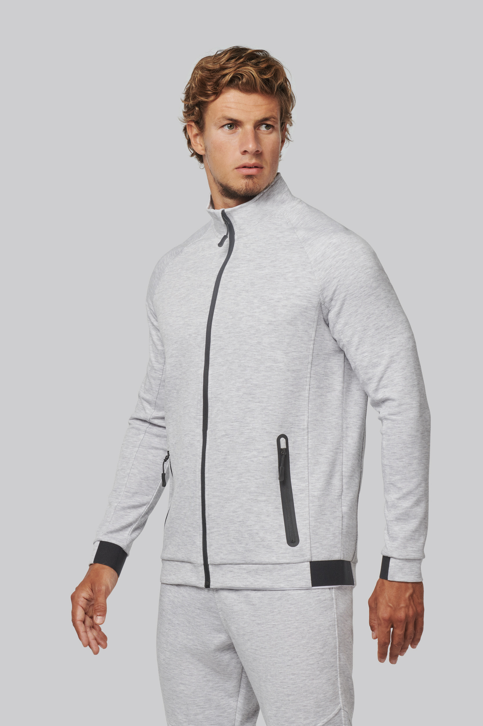 Hombre joven con chándal gris claro de cremallera y bolsillos, fondo neutro.