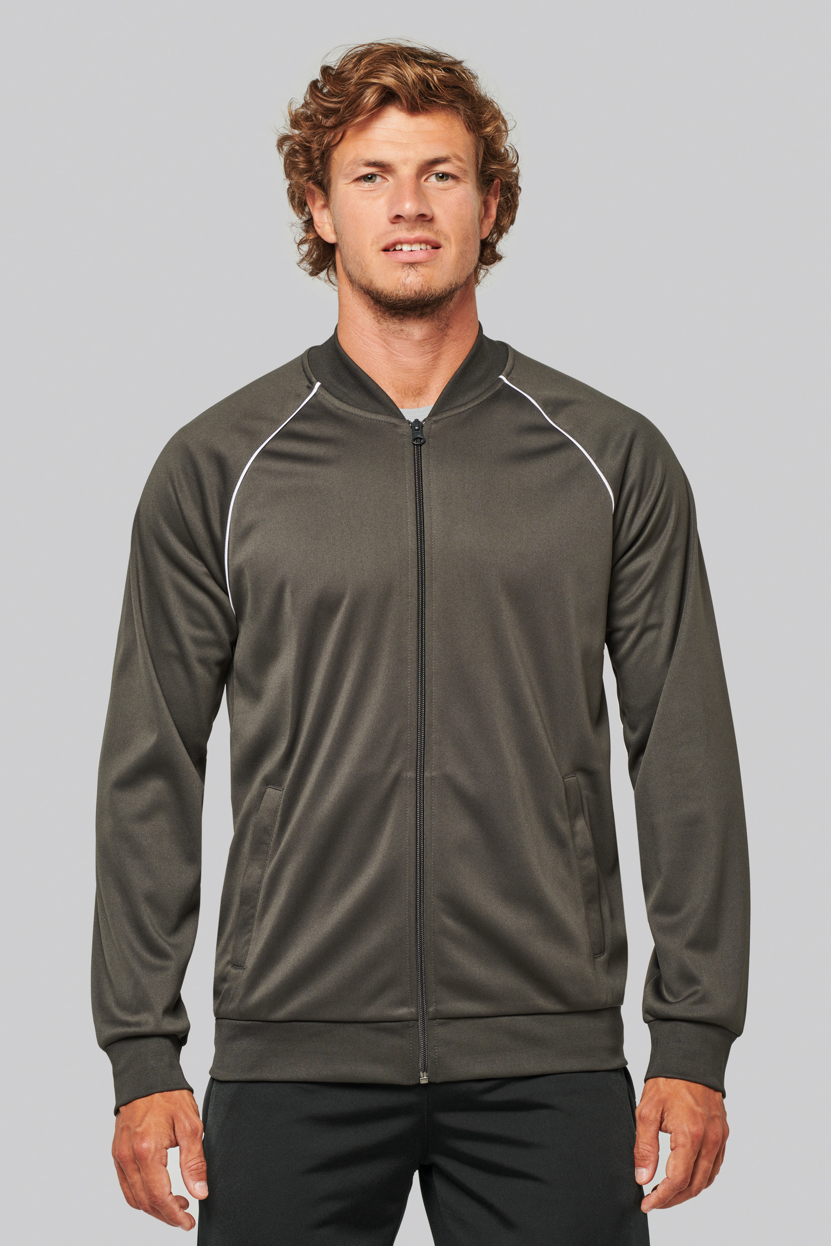 hombre joven de pie con chándal deportivo oscuro y chaqueta con cremallera sobre fondo neutro