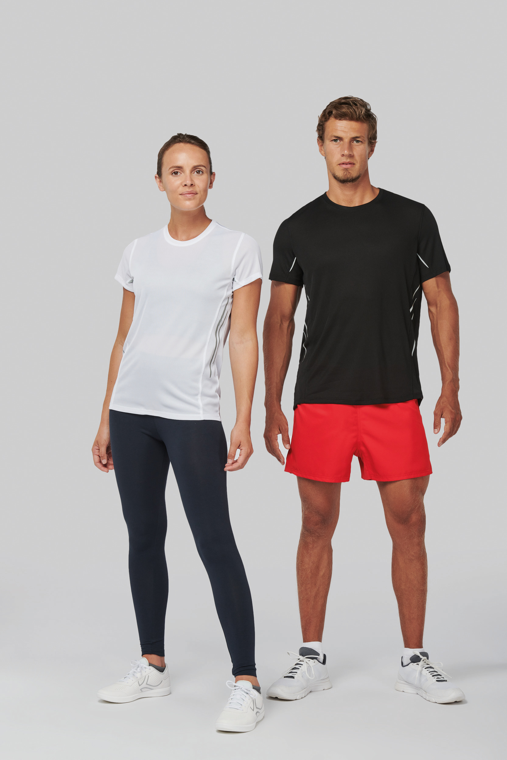 Pareja de modelos deportivos con camiseta y zapatillas blancas, ella con leggings oscuros y él con pantalones cortos rojos sobre fondo neutro.