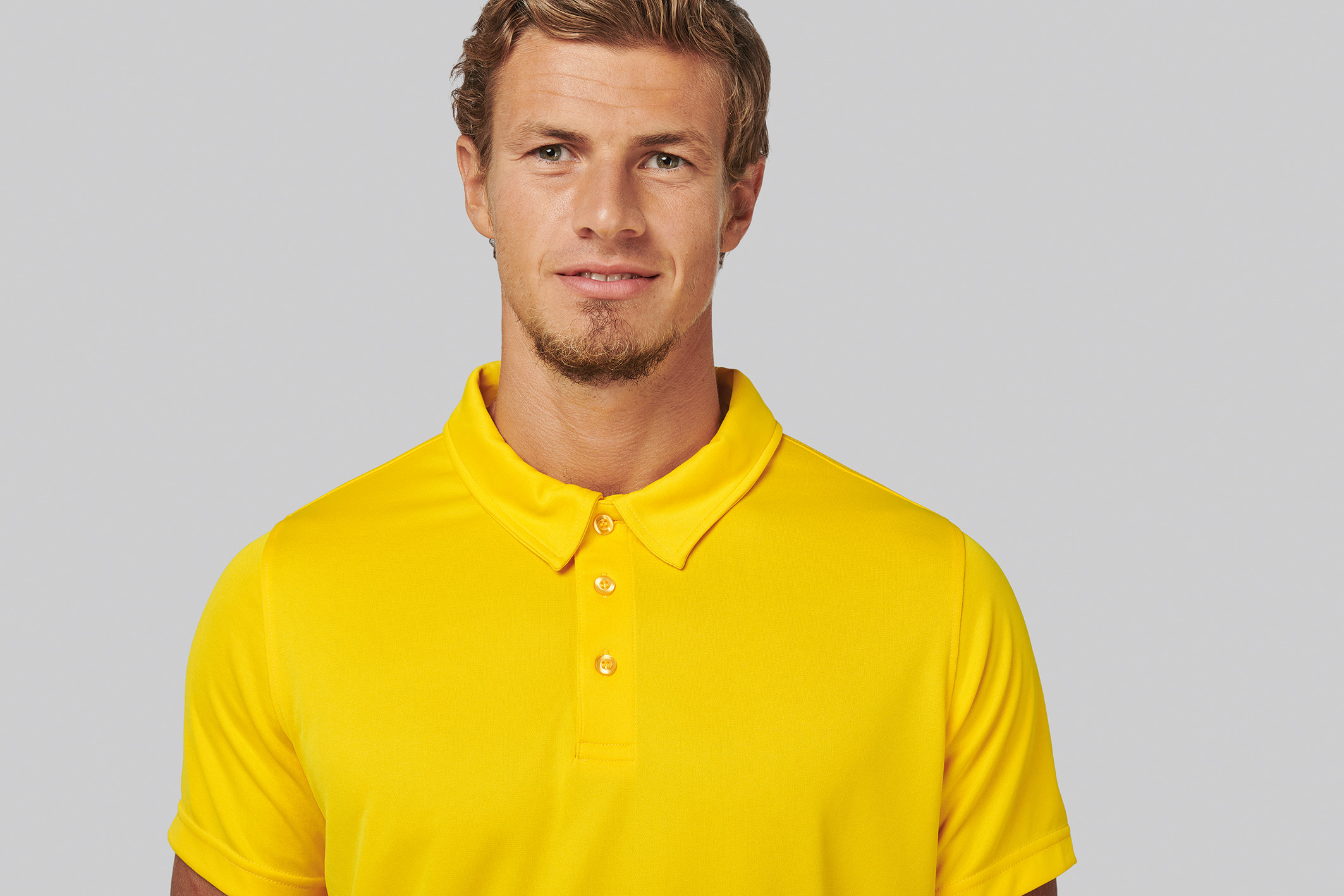 Hombre joven con camiseta polo amarilla sobre fondo neutro.