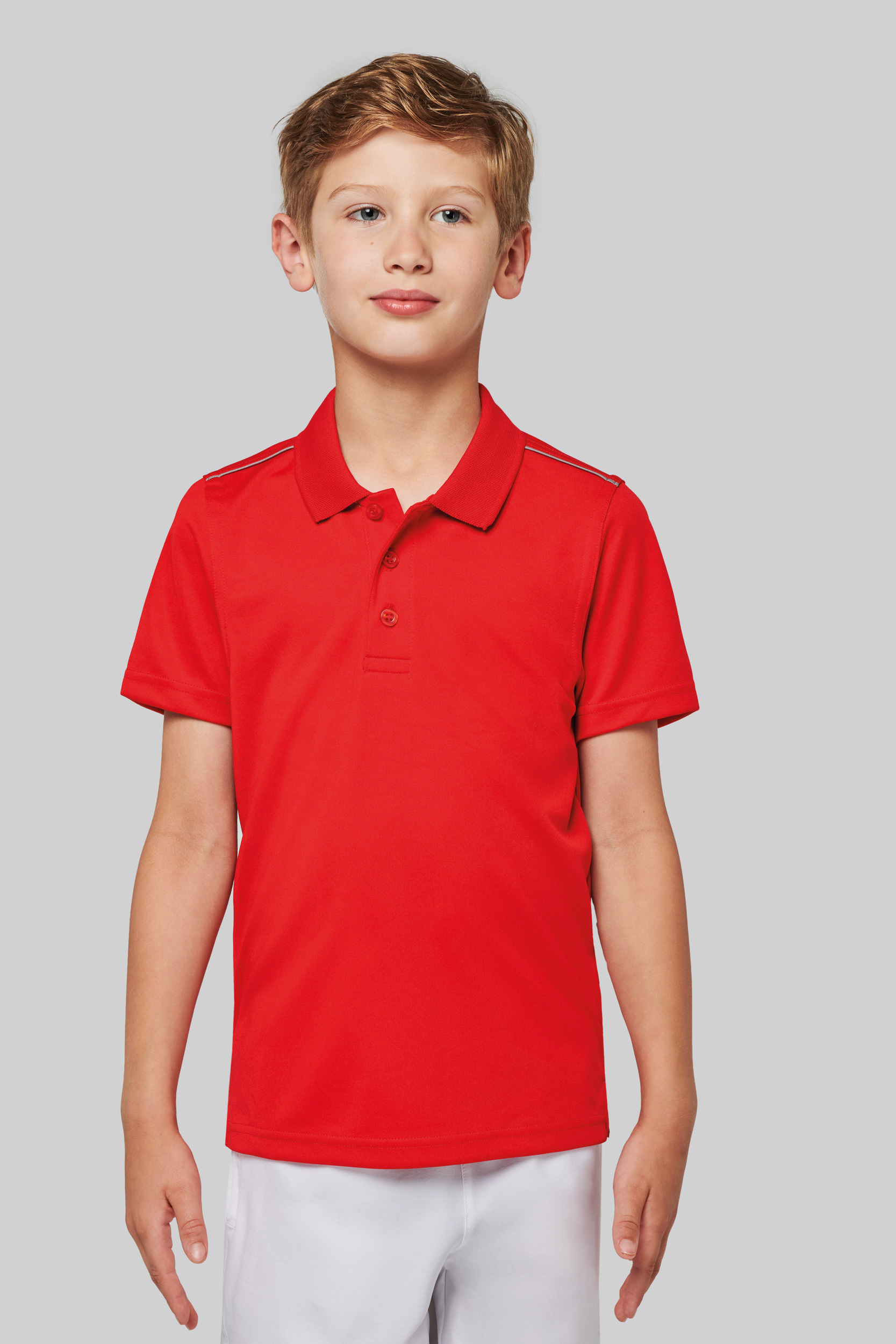 Niño de pie con polo rojo de manga corta sobre fondo gris claro.