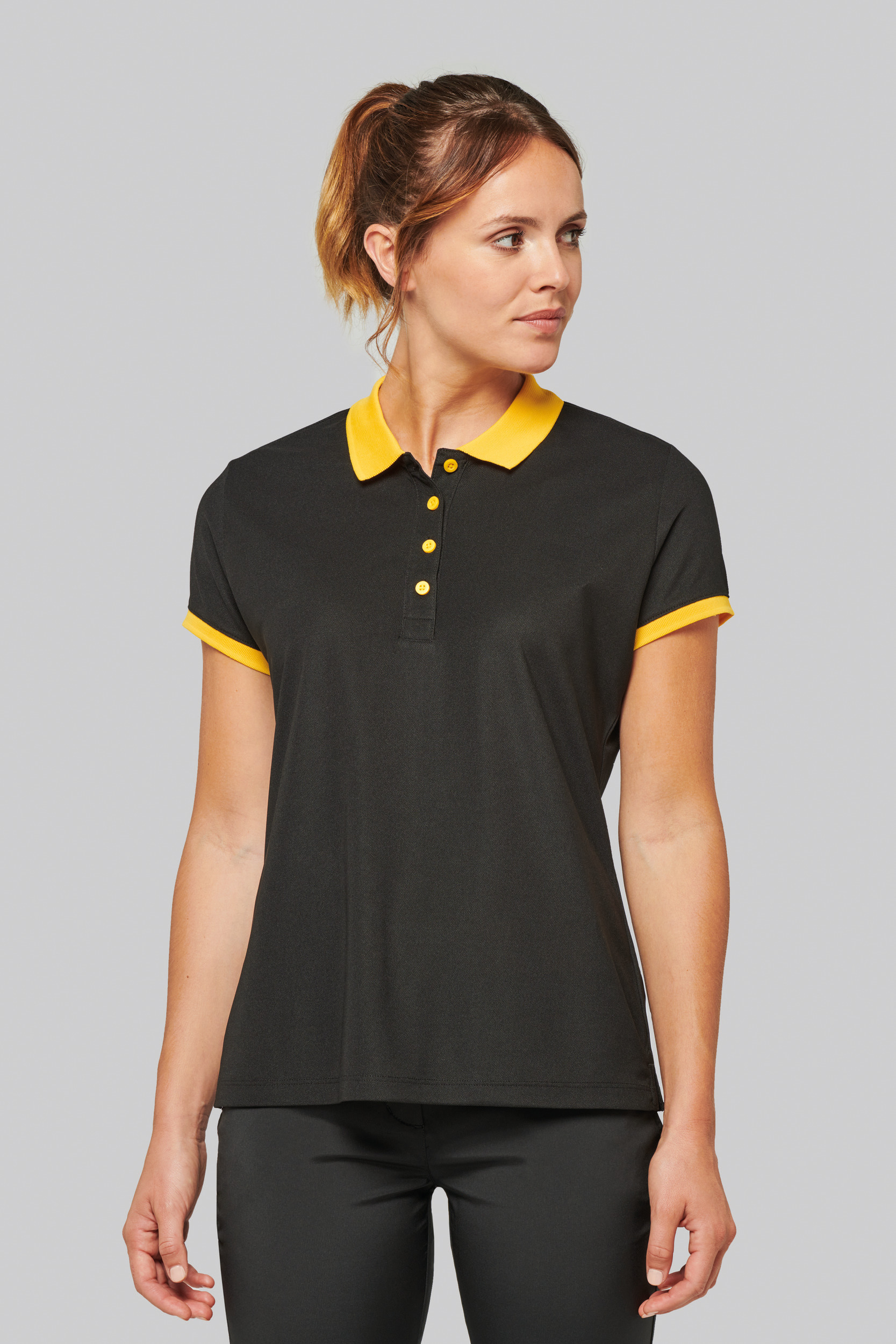 Mujer de pie con polo negro de manga corta y cuello amarillo sobre fondo gris.