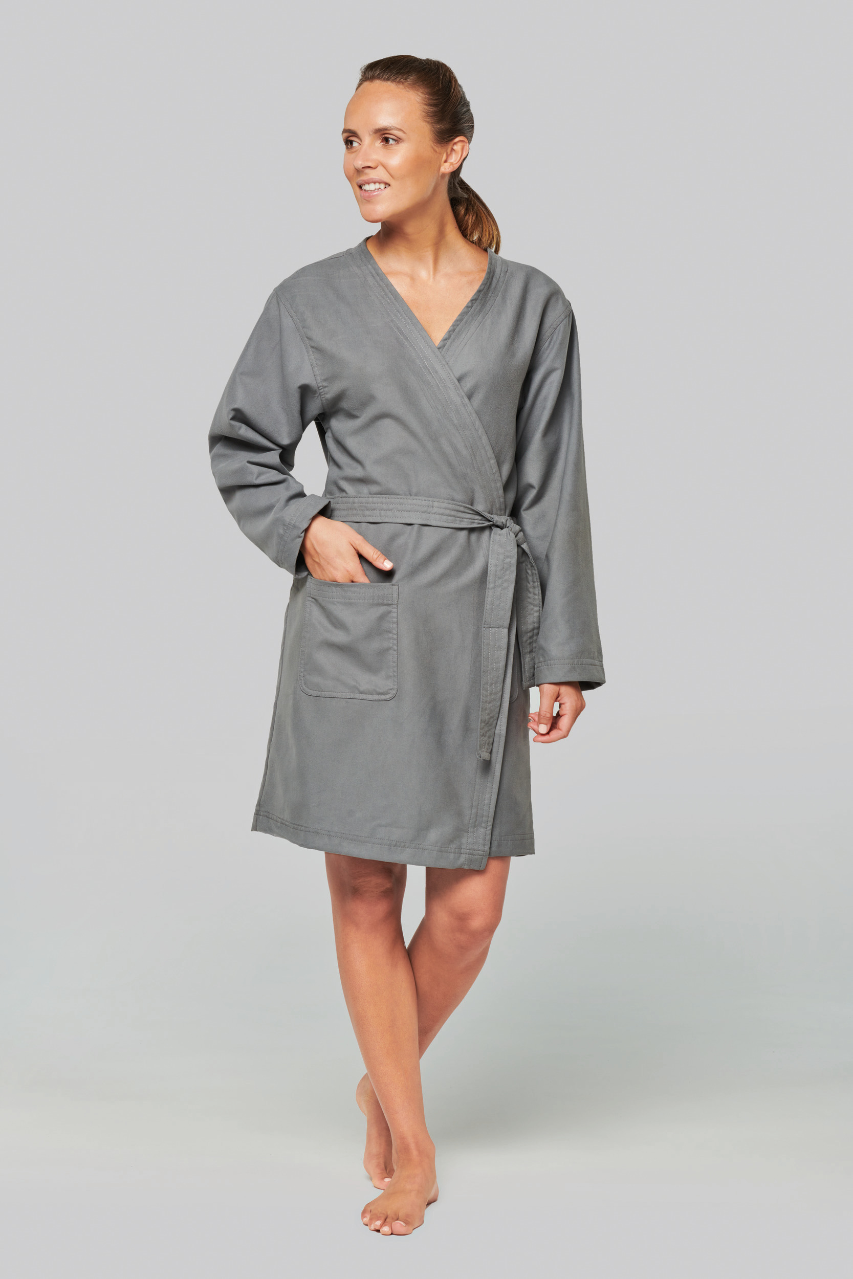 Mujer sonriente de pie con bata gris cruzada y bolsillos en fondo neutro.
