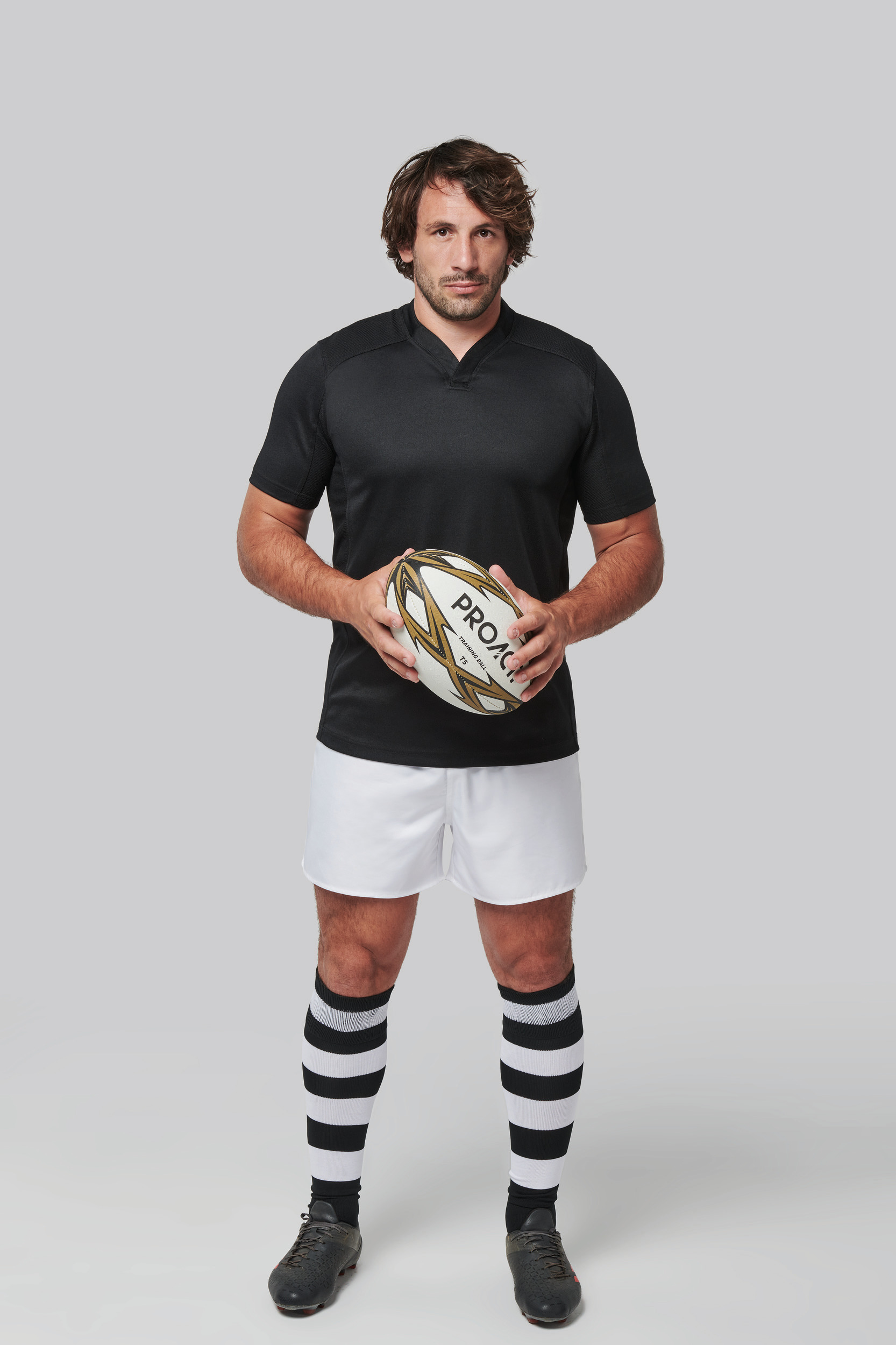 Jugador de rugby vestido con camiseta negra y pantalón blanco sosteniendo un balón sobre fondo neutro.