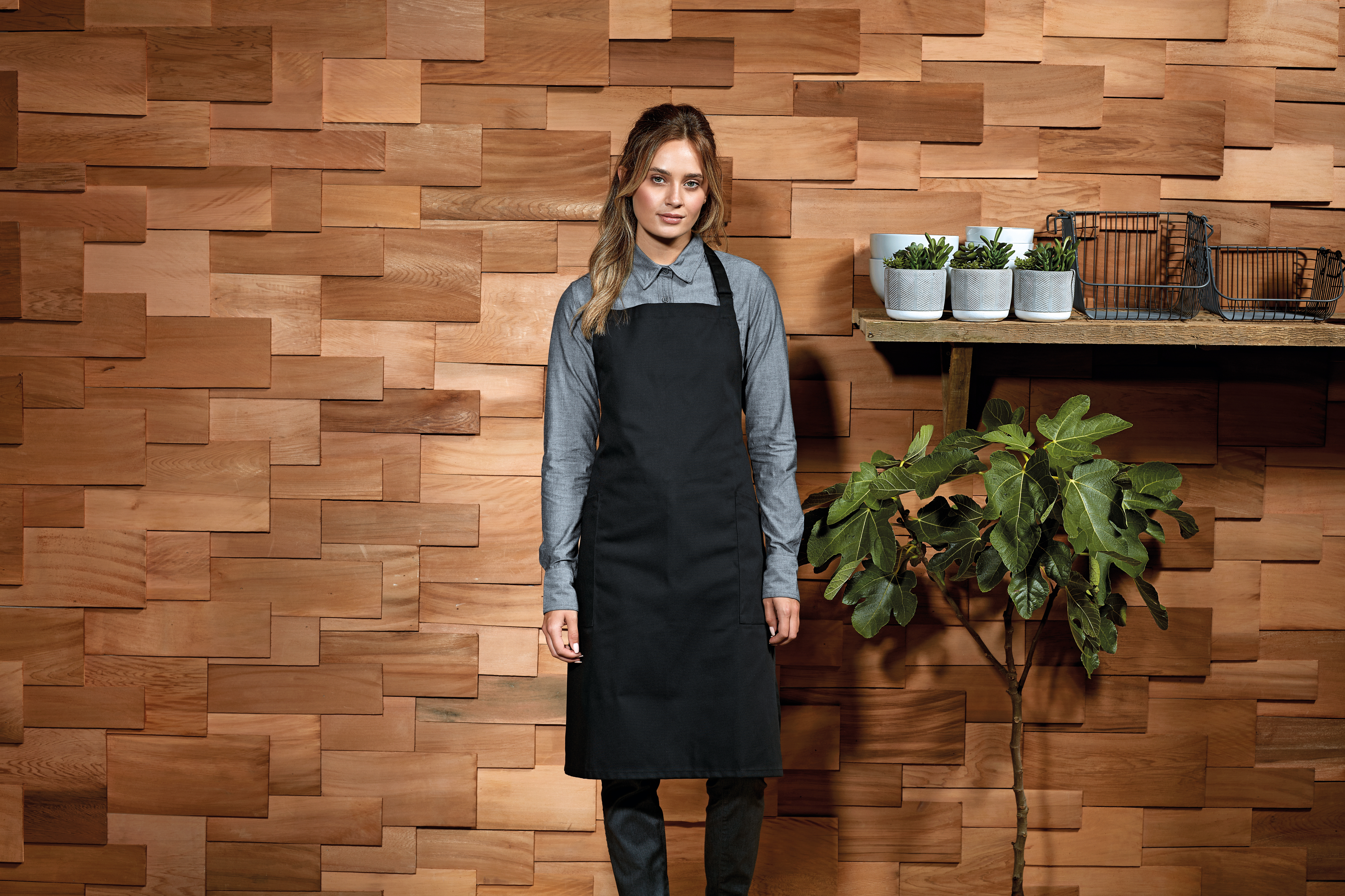 Mujer con delantal negro y camisa gris frente a pared de madera junto a estantería con macetas y planta de interior.