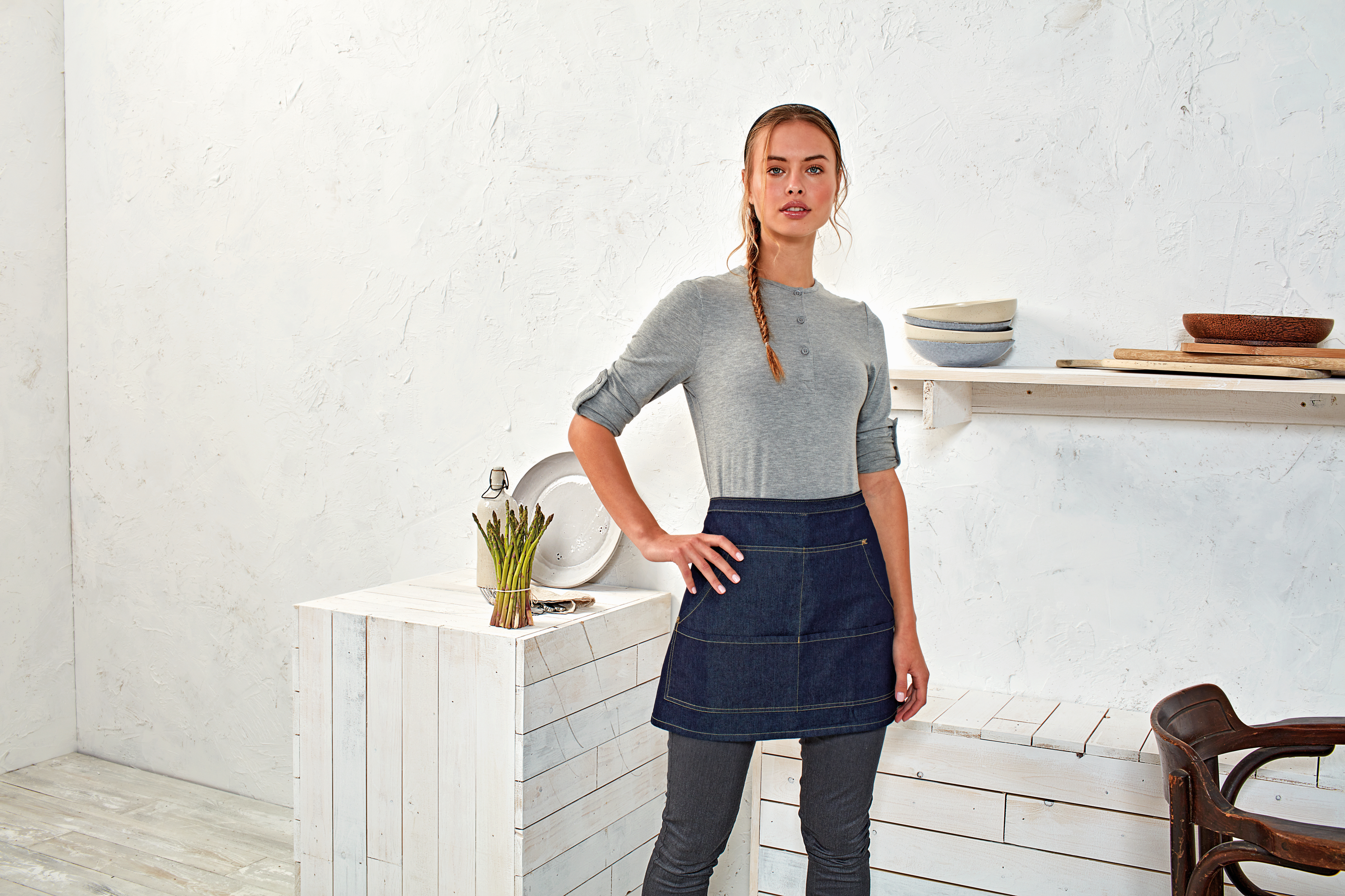 Mujer con delantal vaquero junto a una mesa blanca con espárragos y platos en una cocina de estilo rústico.