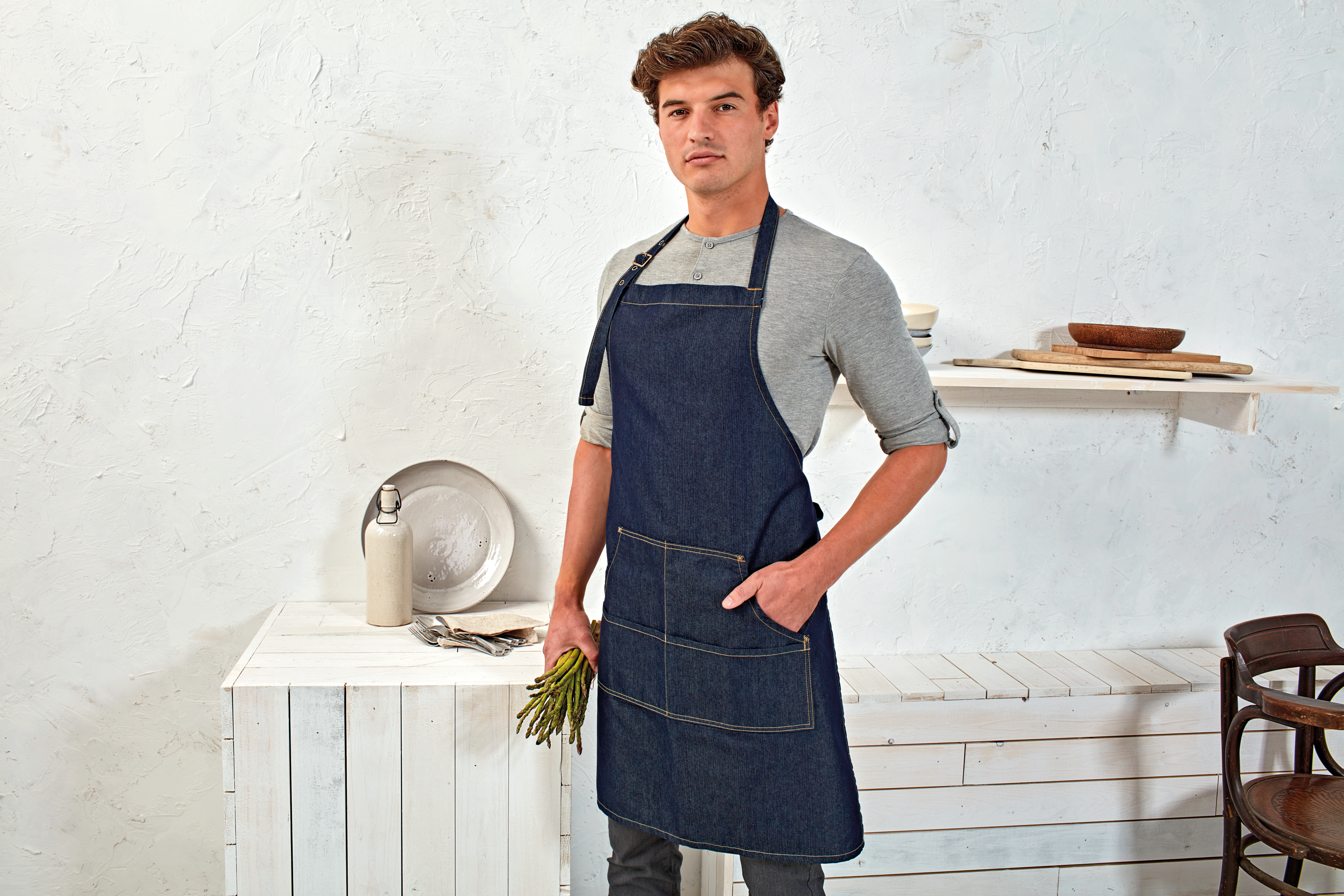 Hombre con delantal vaquero sosteniendo un manojo de espárragos en una cocina de estilo rústico con utensilios y platos sobre madera blanca.