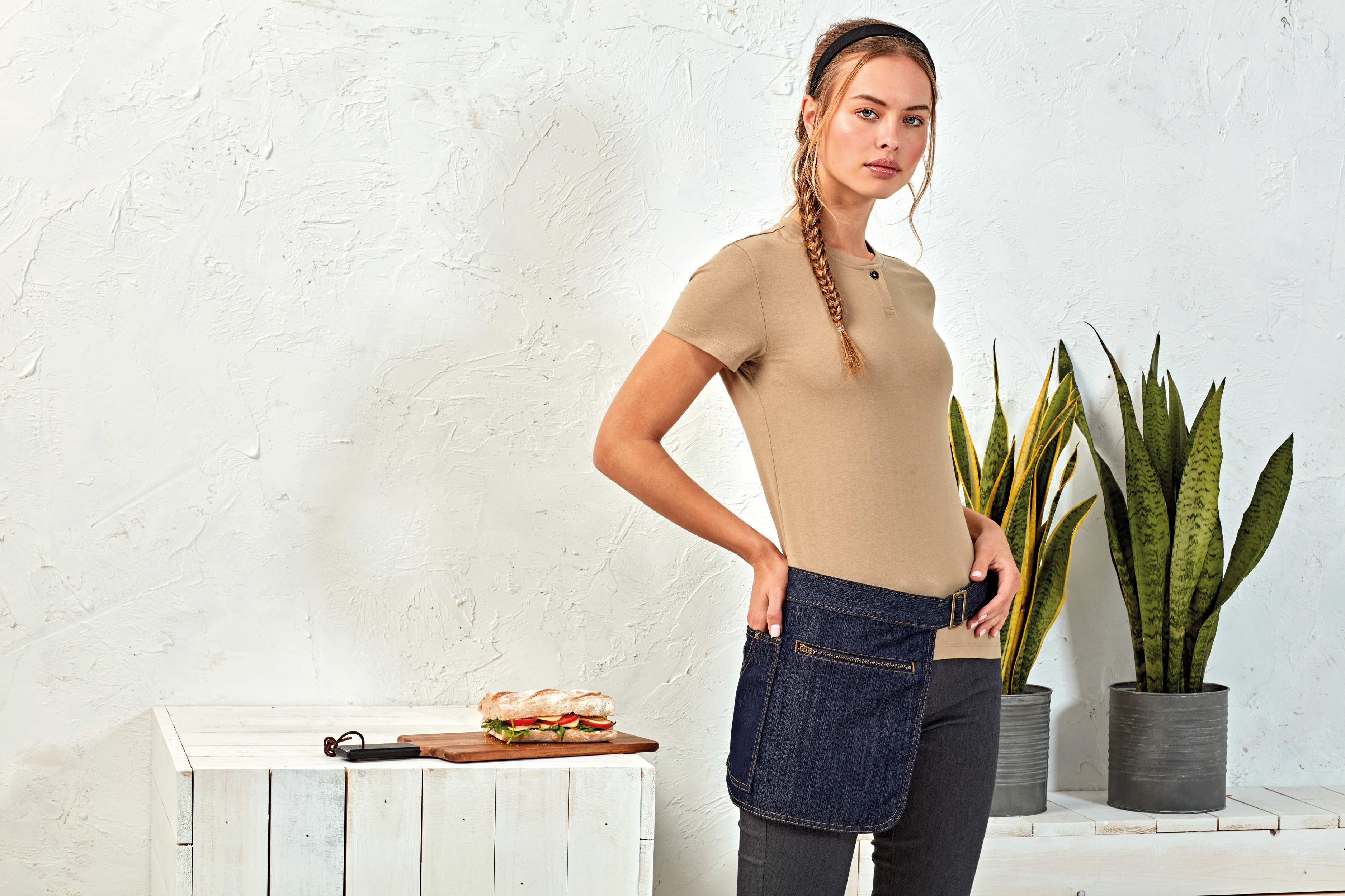 Mujer con media bata vaquera ajustable junto a una mesa con bocadillo y plantas domésticas
