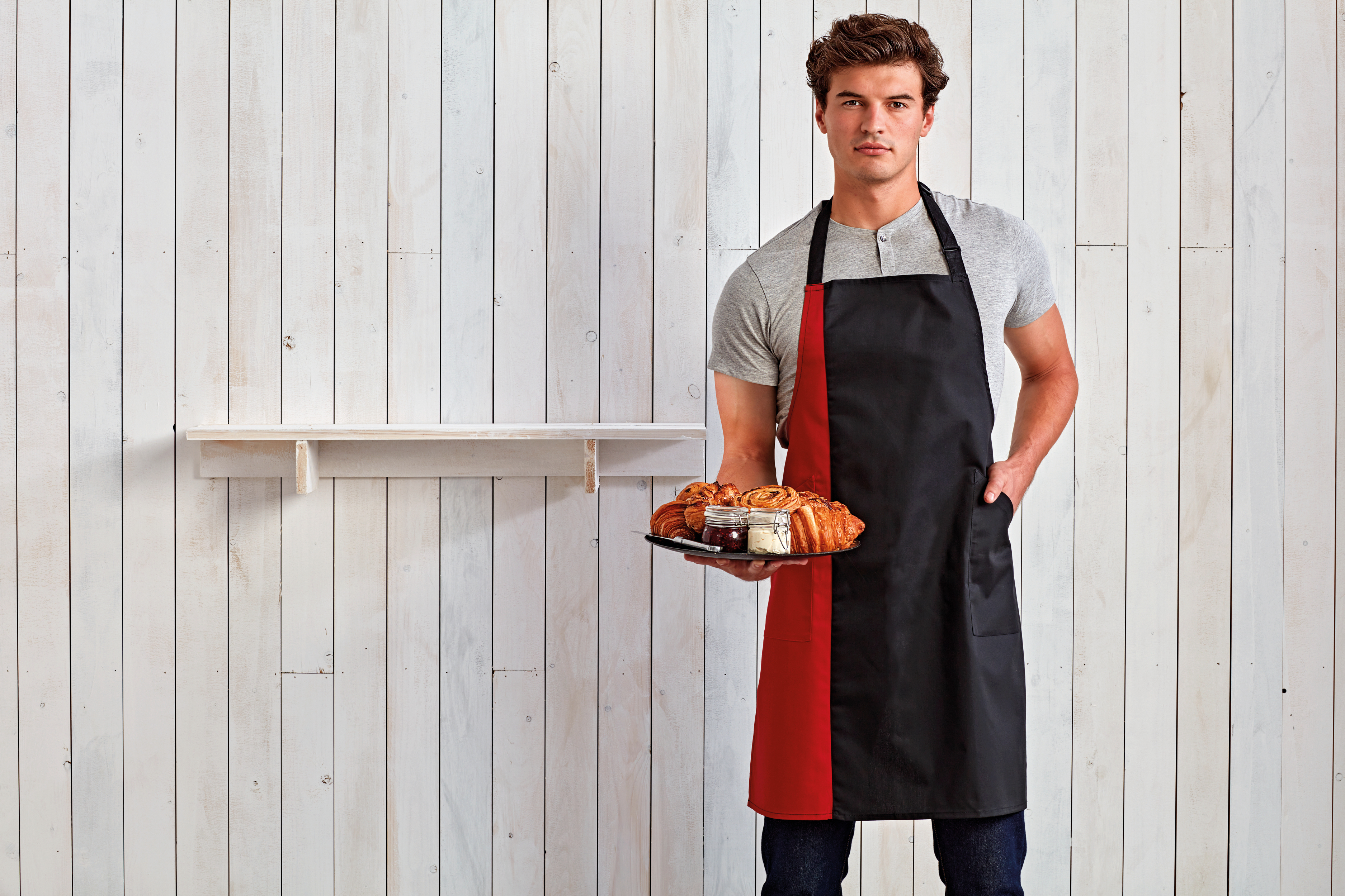 Hombre con delantal negro y rojo sosteniendo una bandeja de croissants y mermelada frente a una pared de tablones de madera clara