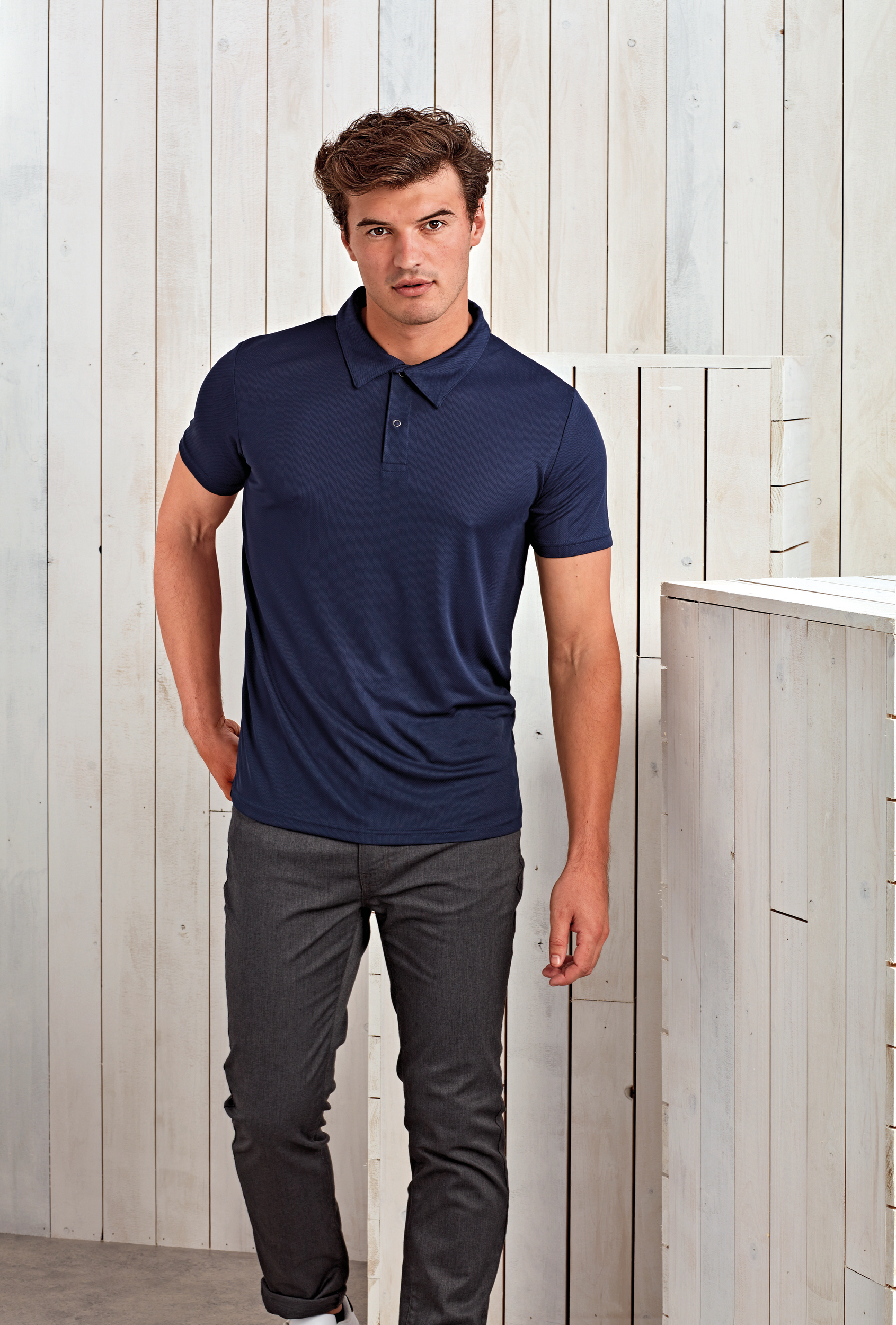 Hombre joven de pie con polo azul marino y pantalones grises frente a un fondo de listones de madera blanca