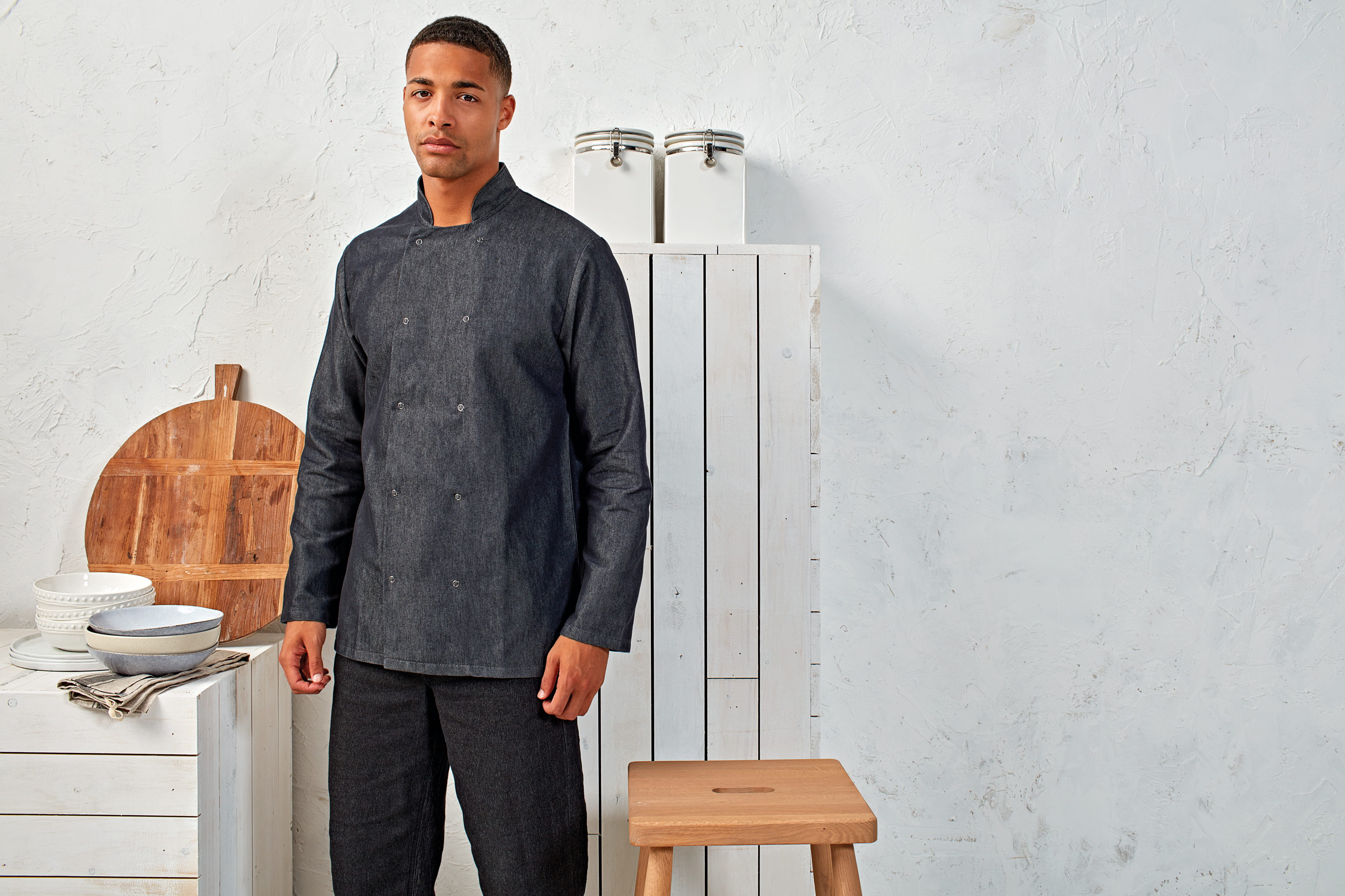 Hombre con chaqueta de chef de mezclilla oscura junto a tabla de cortar, cuencos y taburete en cocina minimalista