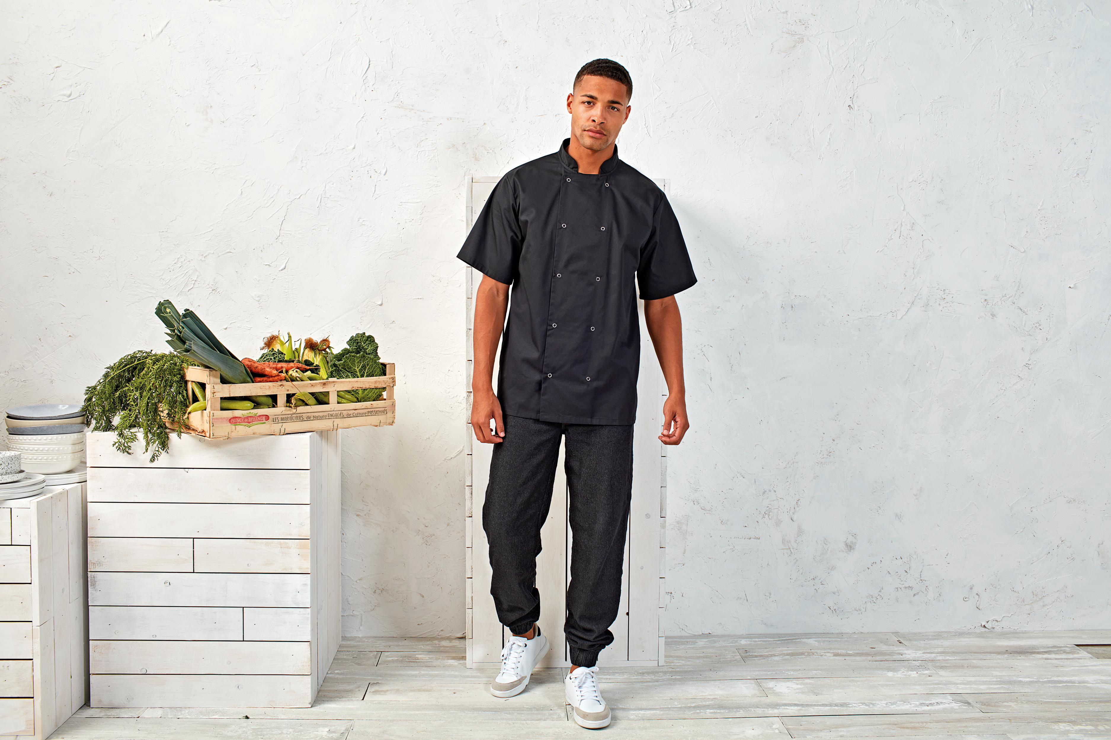 Chef joven con chaquetilla negra junto a caja de verduras frescas sobre cajones de madera blanca.