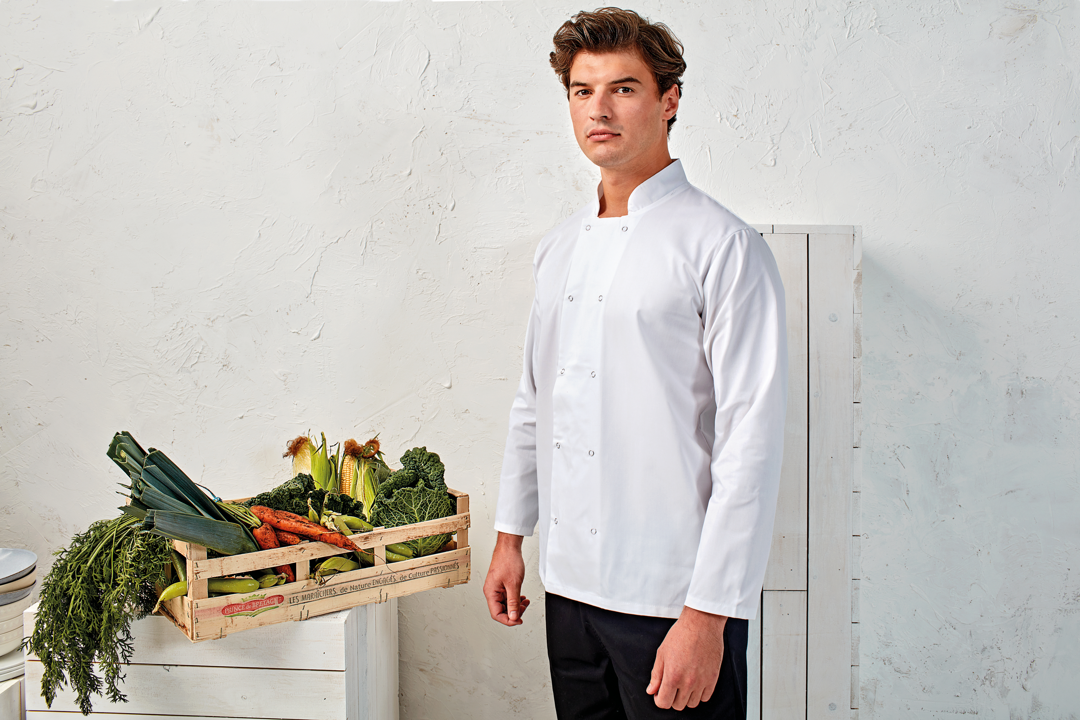 chef con chaqueta blanca de pie junto a una caja de verduras frescas sobre un mueble blanco