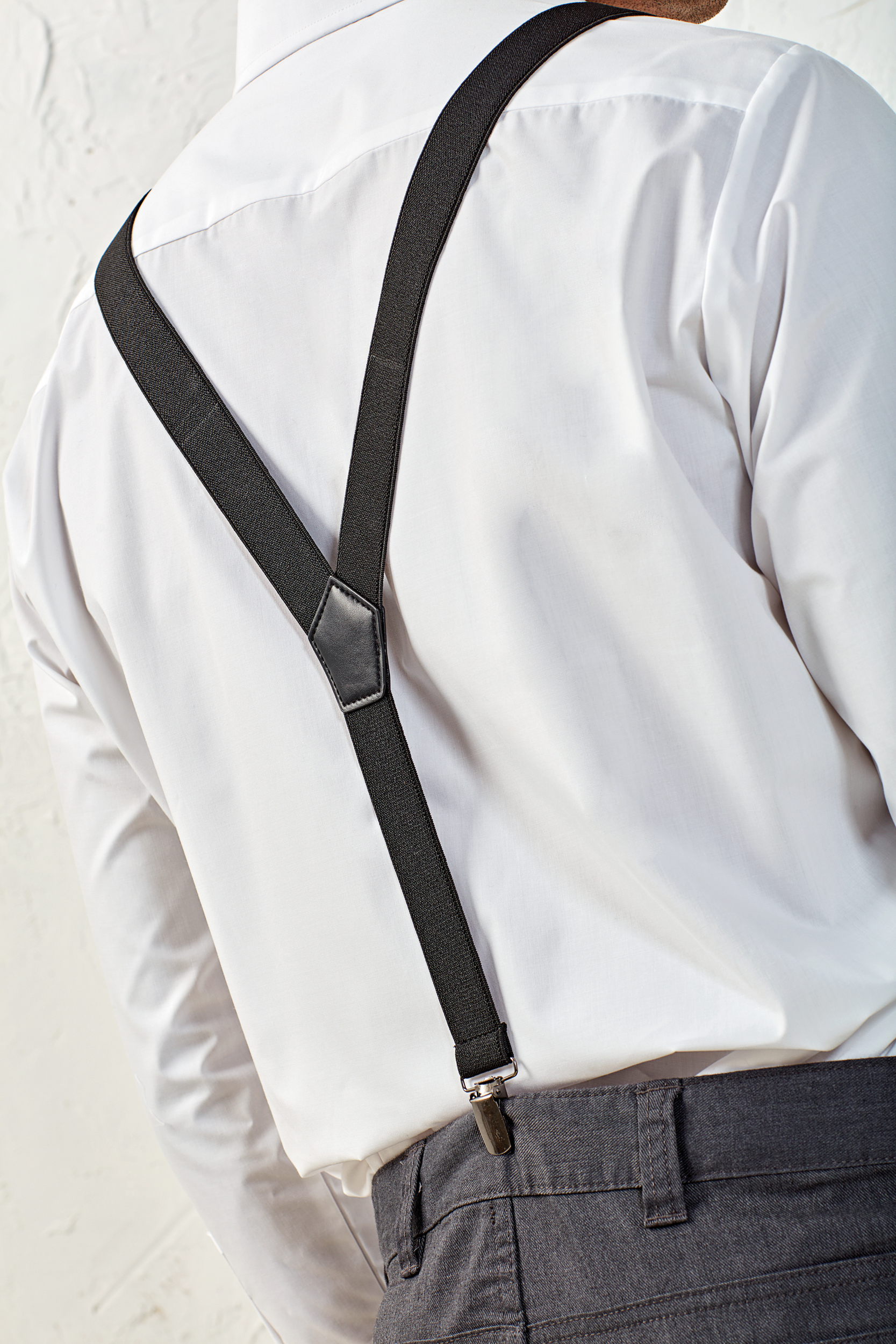 Hombre de espaldas con camisa blanca y tirantes negros cruzados sujetando pantalón gris.