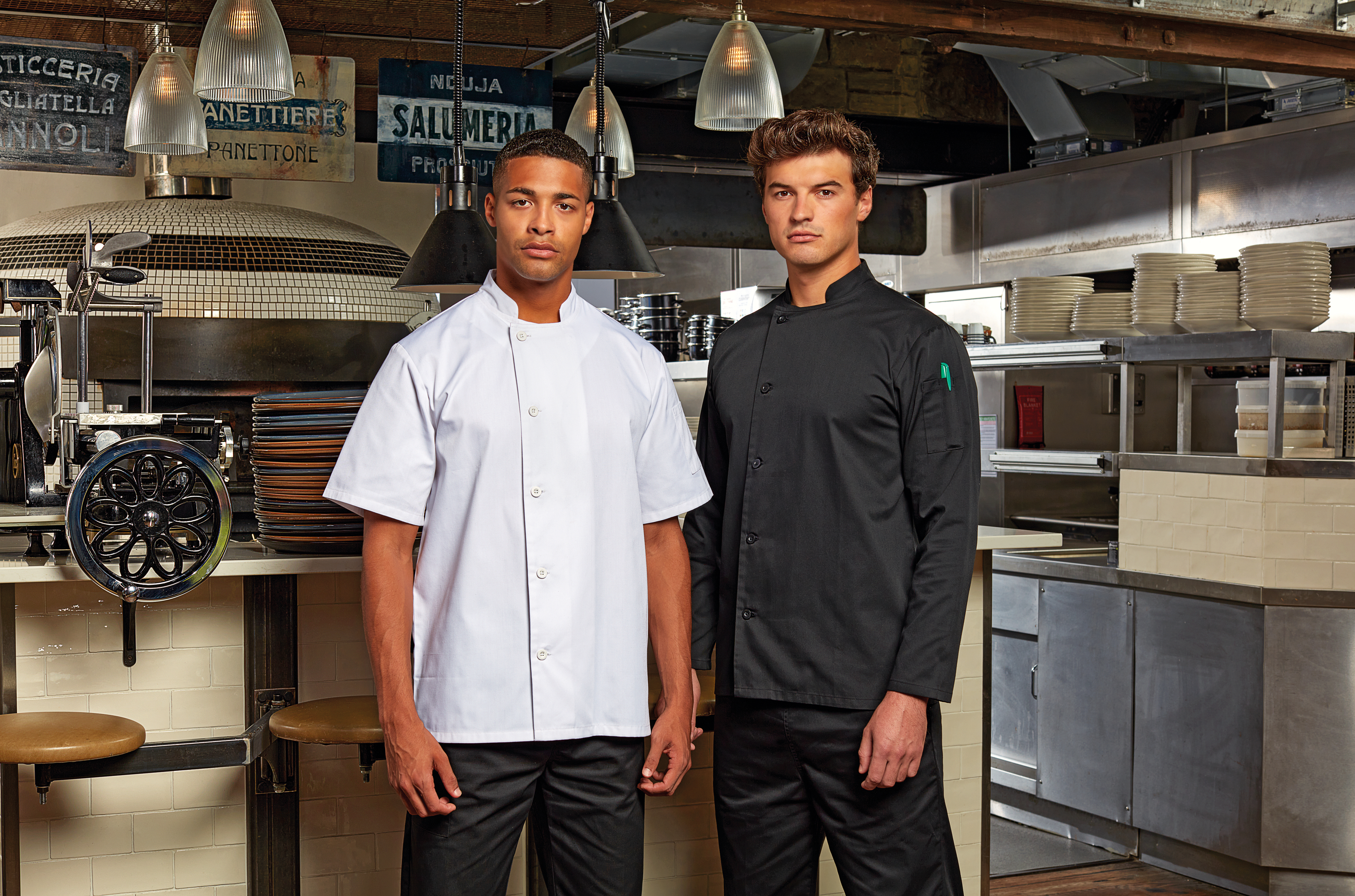 Dos cocineros de pie en una cocina profesional junto a un horno de leña y una máquina cortadora de embutidos.