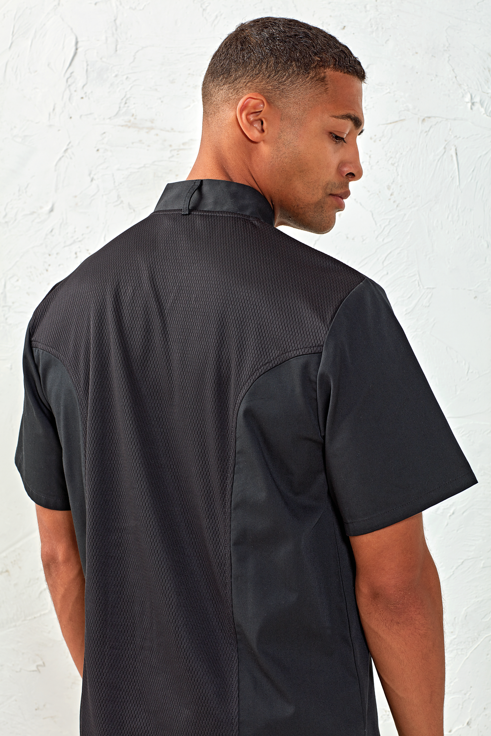 Hombre de espaldas con camisa negra de manga corta y panel trasero de tejido transpirable frente a pared blanca