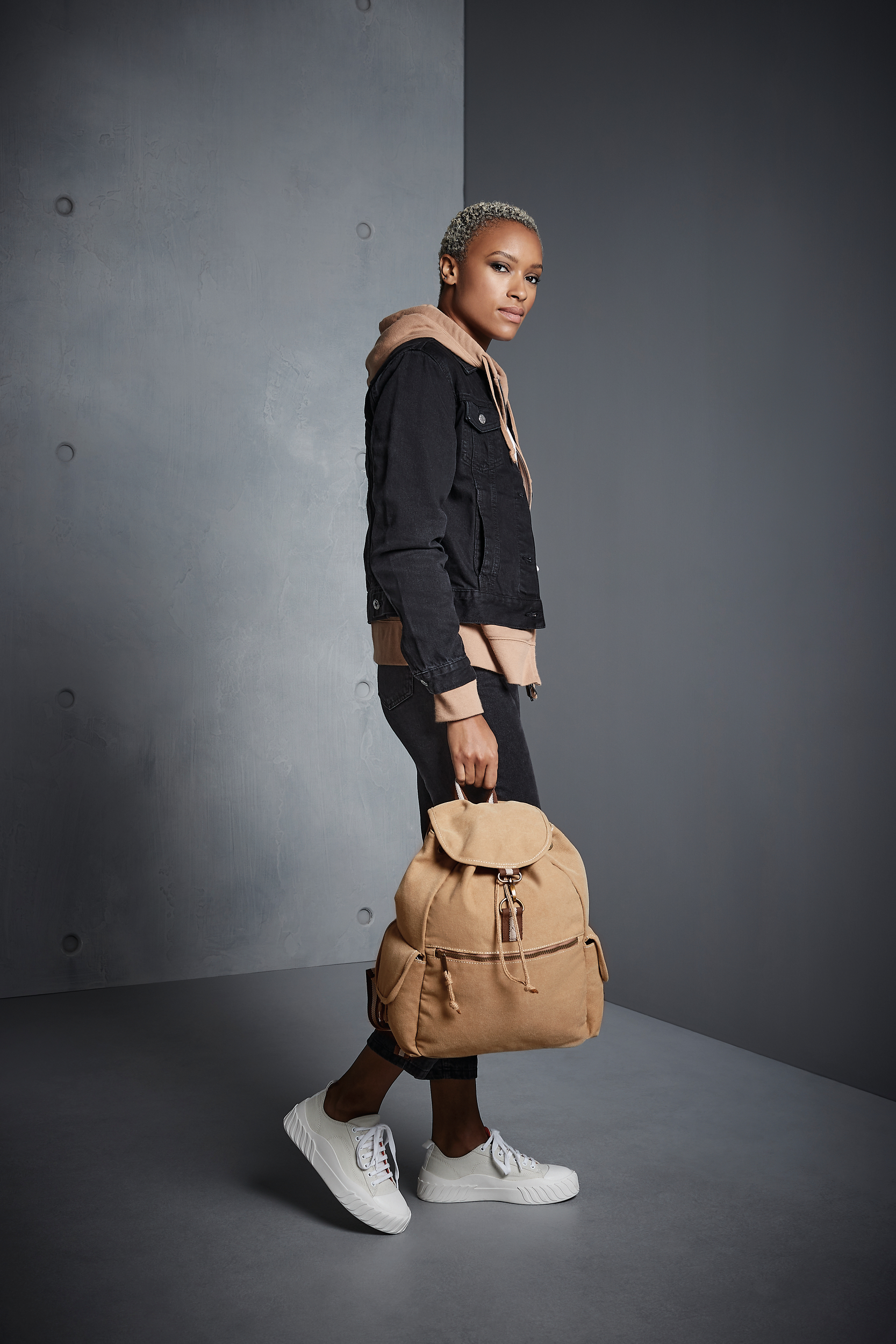 Mujer joven de pie con mochila de tela beige y zapatillas blancas sobre fondo gris.