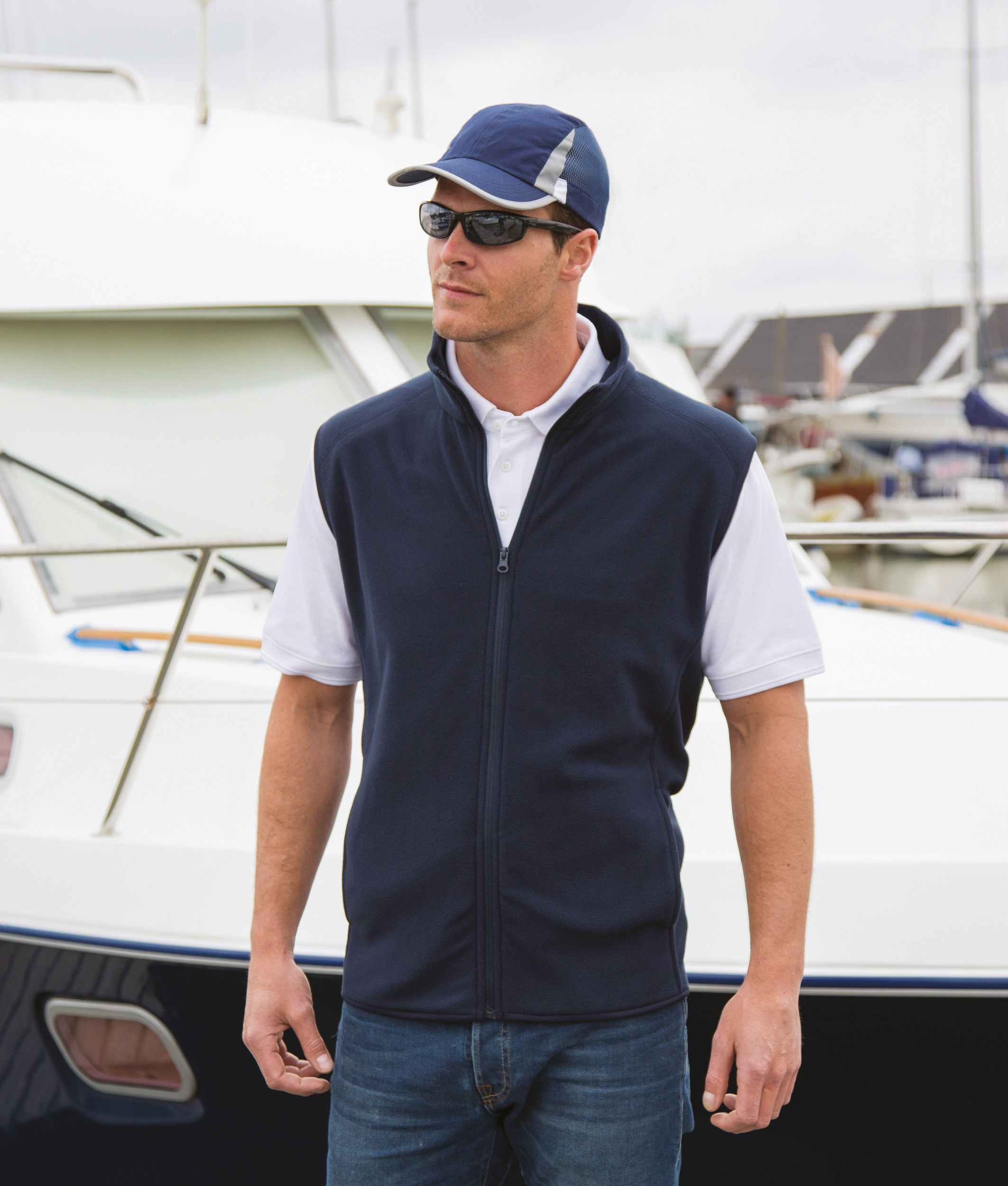 hombre con chaleco azul marino y gorra junto a un barco en el puerto