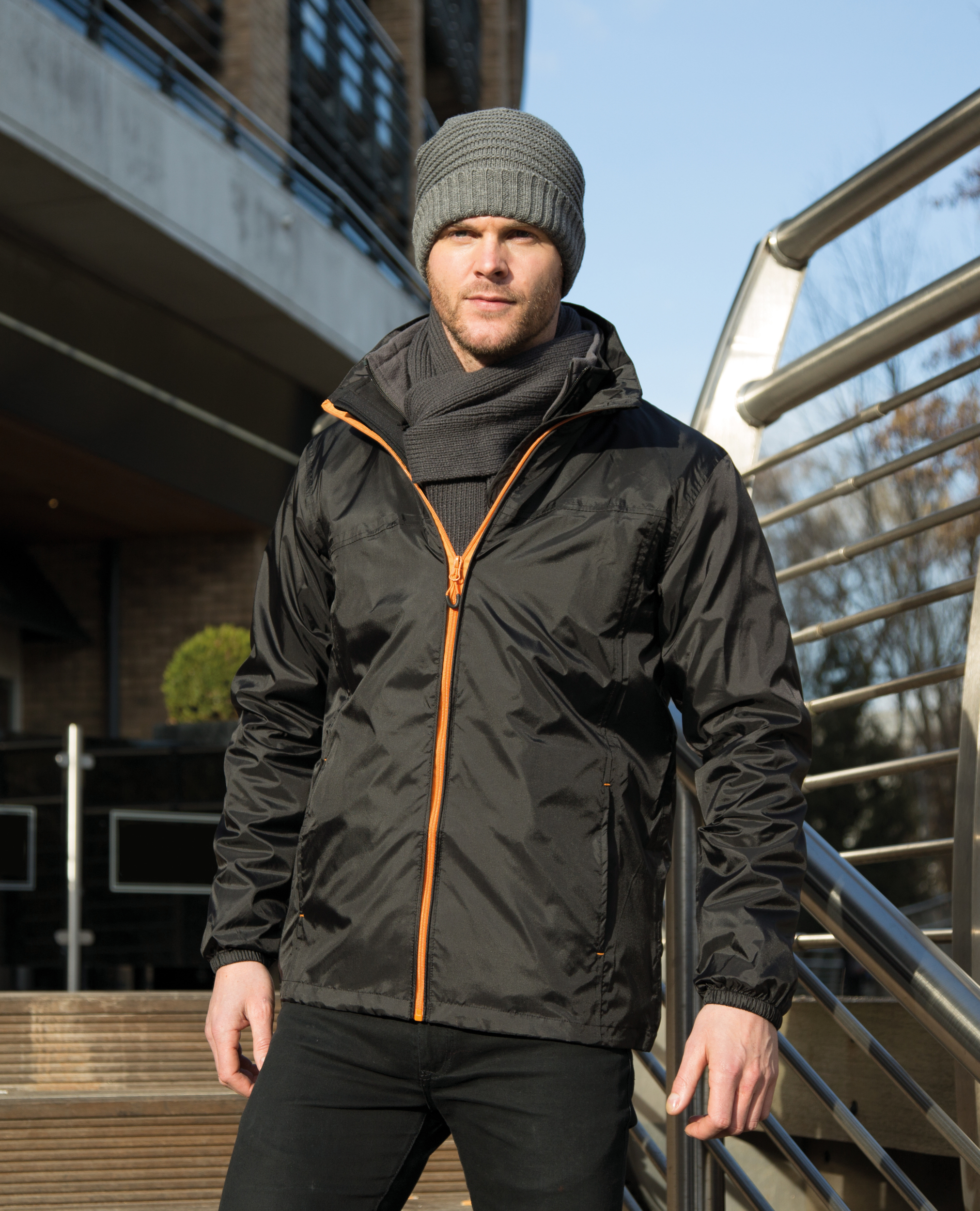 Hombre con chaqueta negra, gorro y bufanda de invierno apoyado en una barandilla urbana.