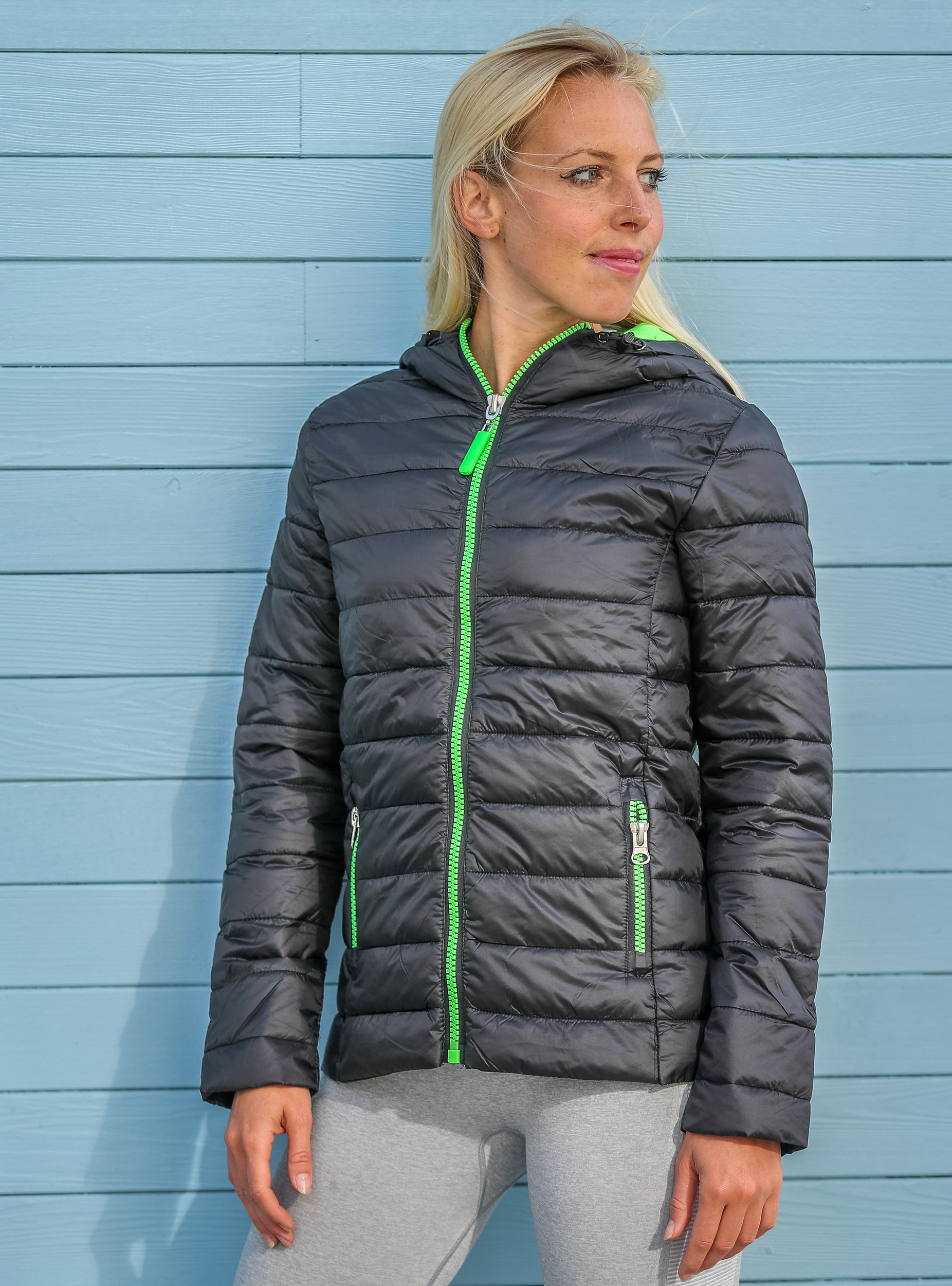 Mujer con chaqueta acolchada negra y cremalleras verdes frente a una pared de madera azul