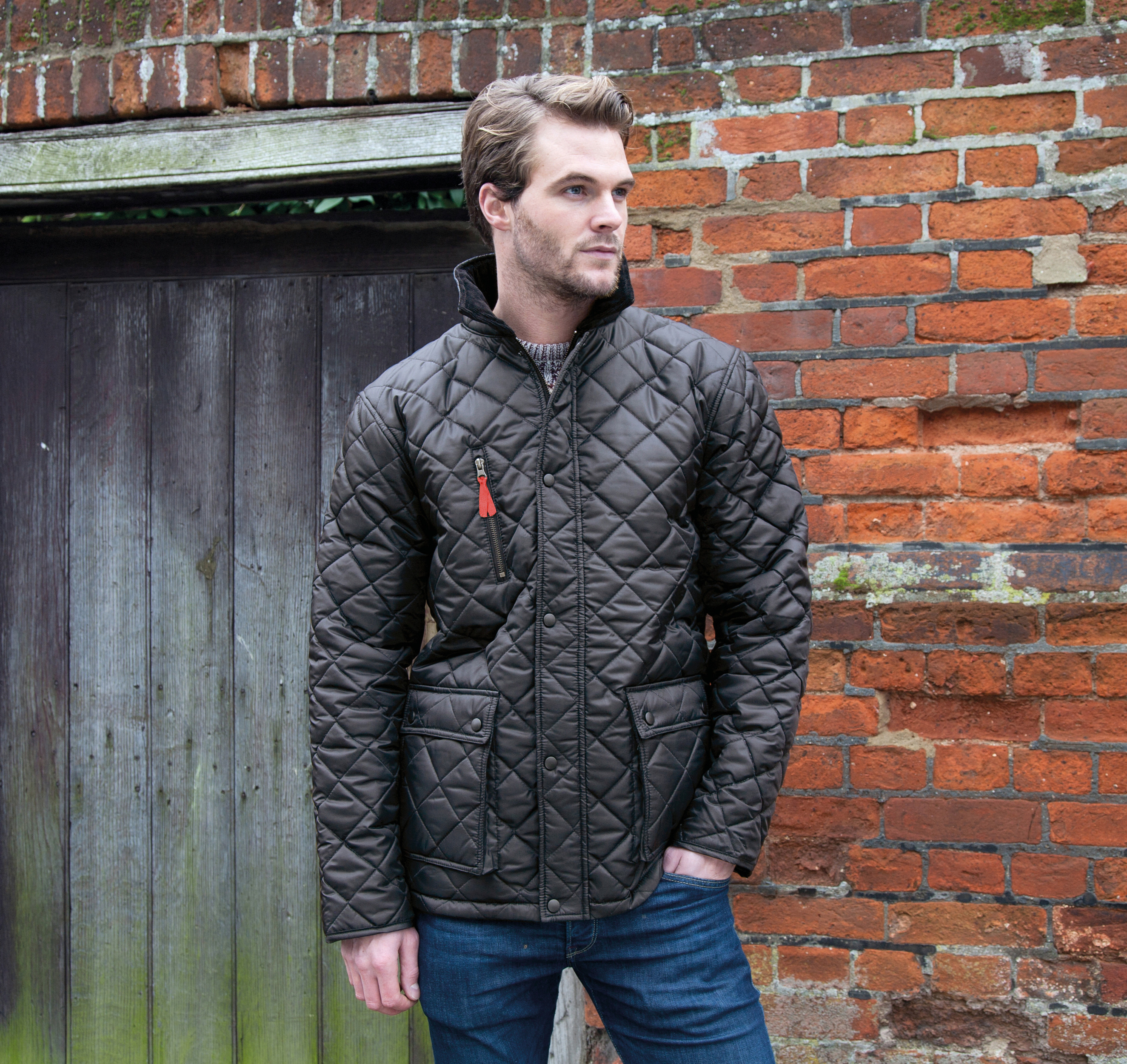 Hombre vestido con chaqueta acolchada oscura apoyado junto a un muro de ladrillo y una puerta de madera envejecida