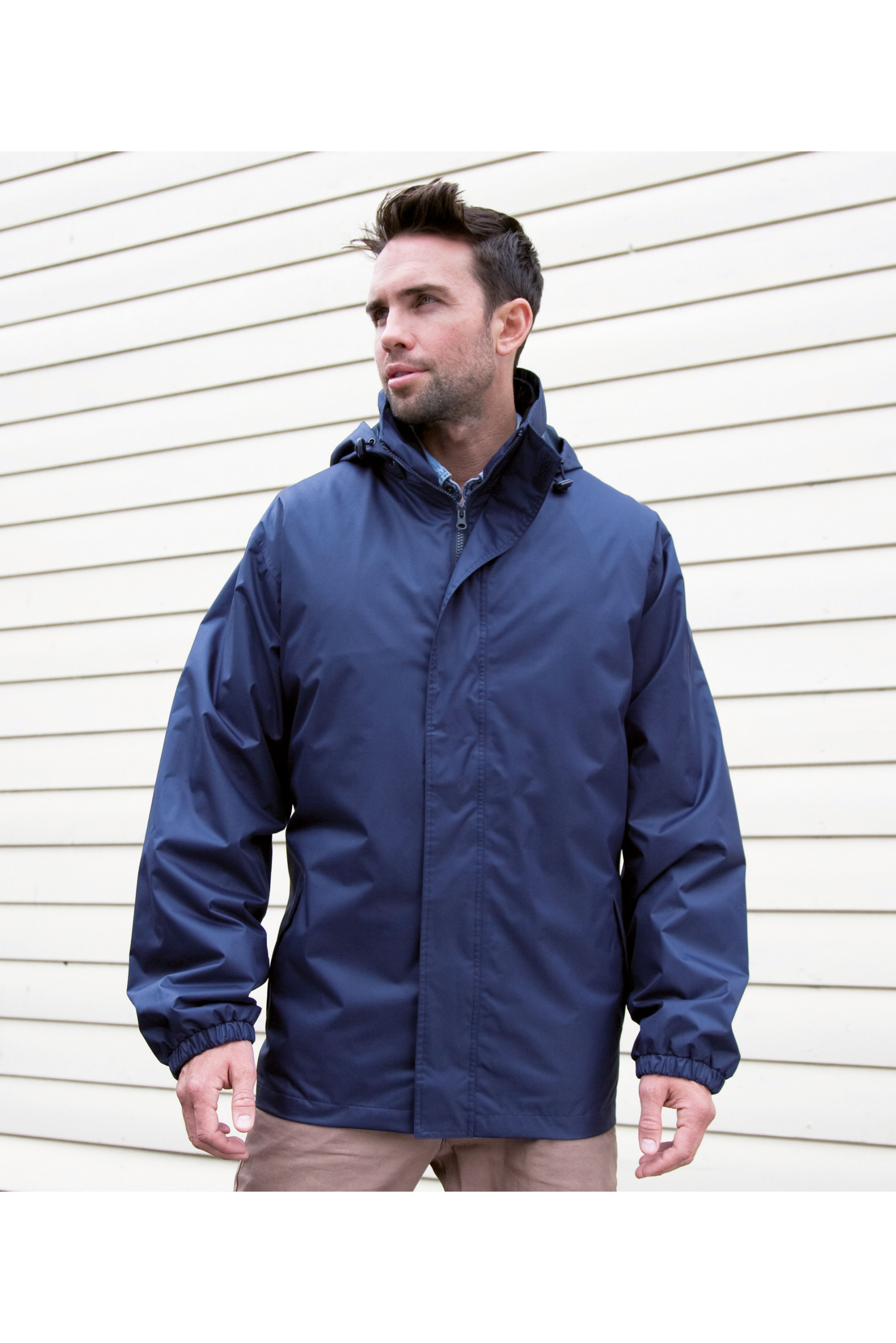 Hombre con chaqueta azul y capucha de lluvia junto a una fachada de listones blancos.