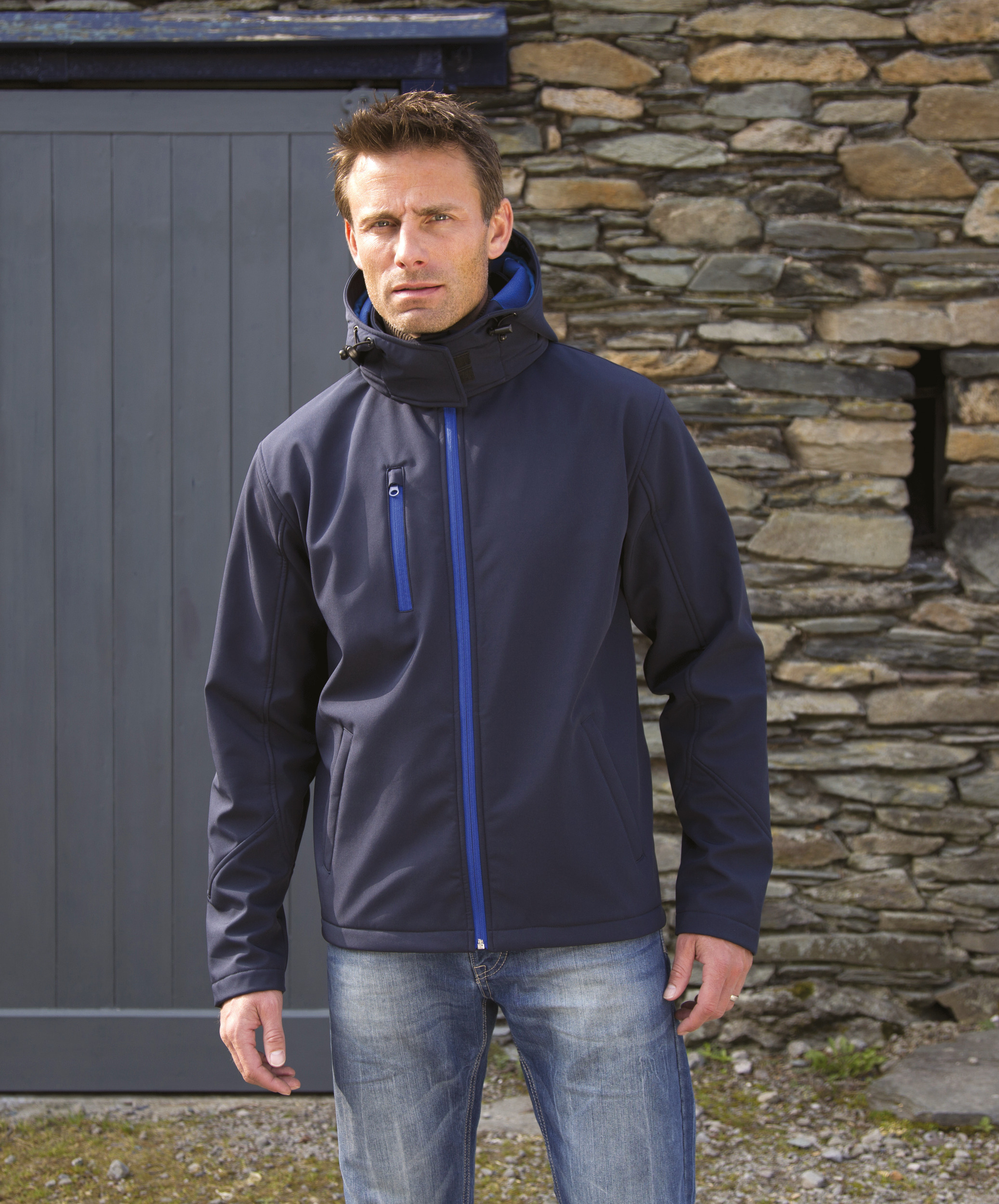 Hombre vistiendo chaqueta softshell azul con capucha frente a un muro de piedra